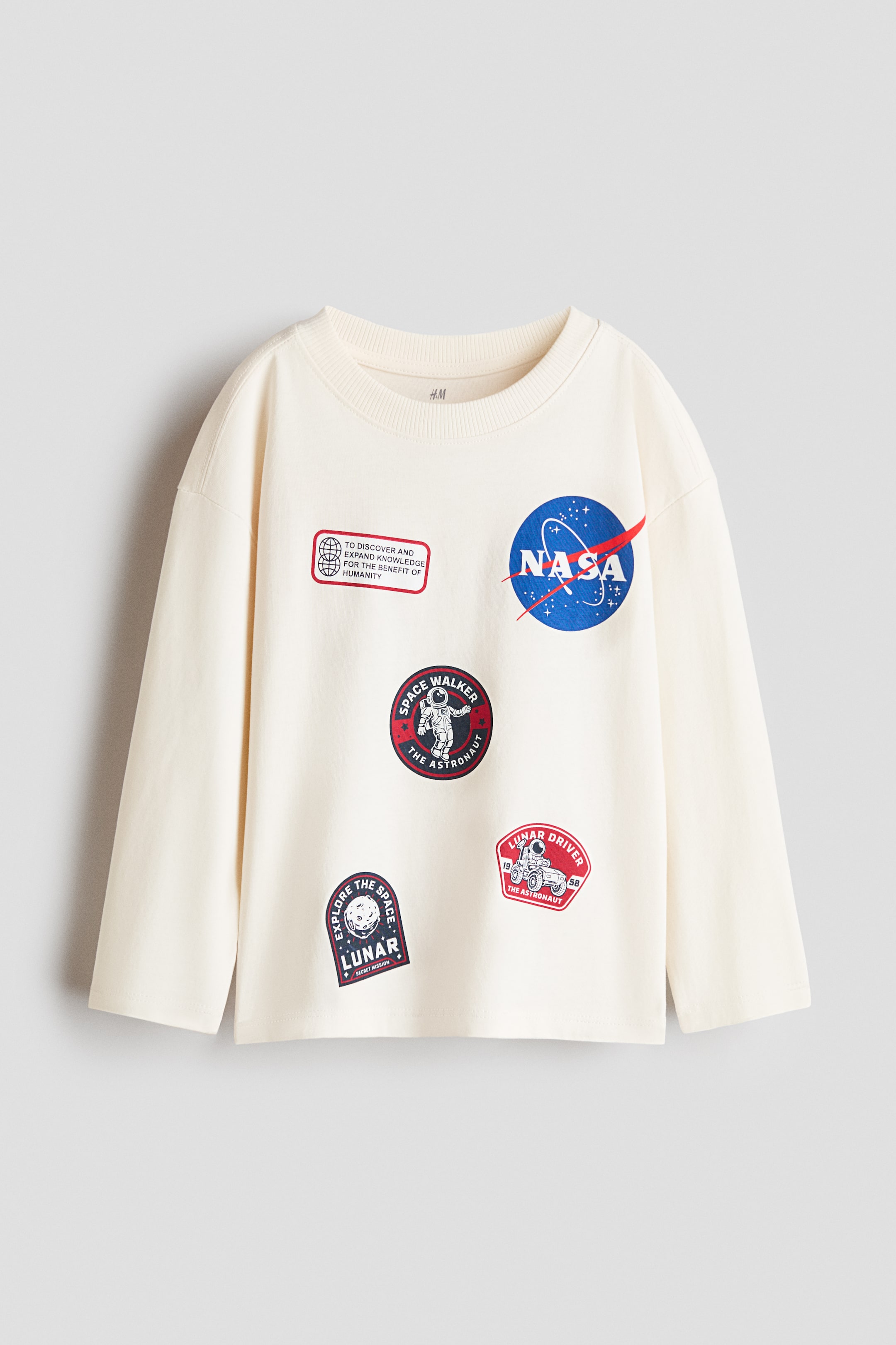 Agrandir l'image: Vue de face d’un t-shirt manches longues écru en jersey de coton doux, doté d’un col rond côtelé, d’épaules tombantes et d’un ourlet droit. Le devant présente plusieurs motifs graphiques imprimés sur le thème de la NASA, dont l’emblématique logo NASA bleu et विविध badges d’exploration spatiale.