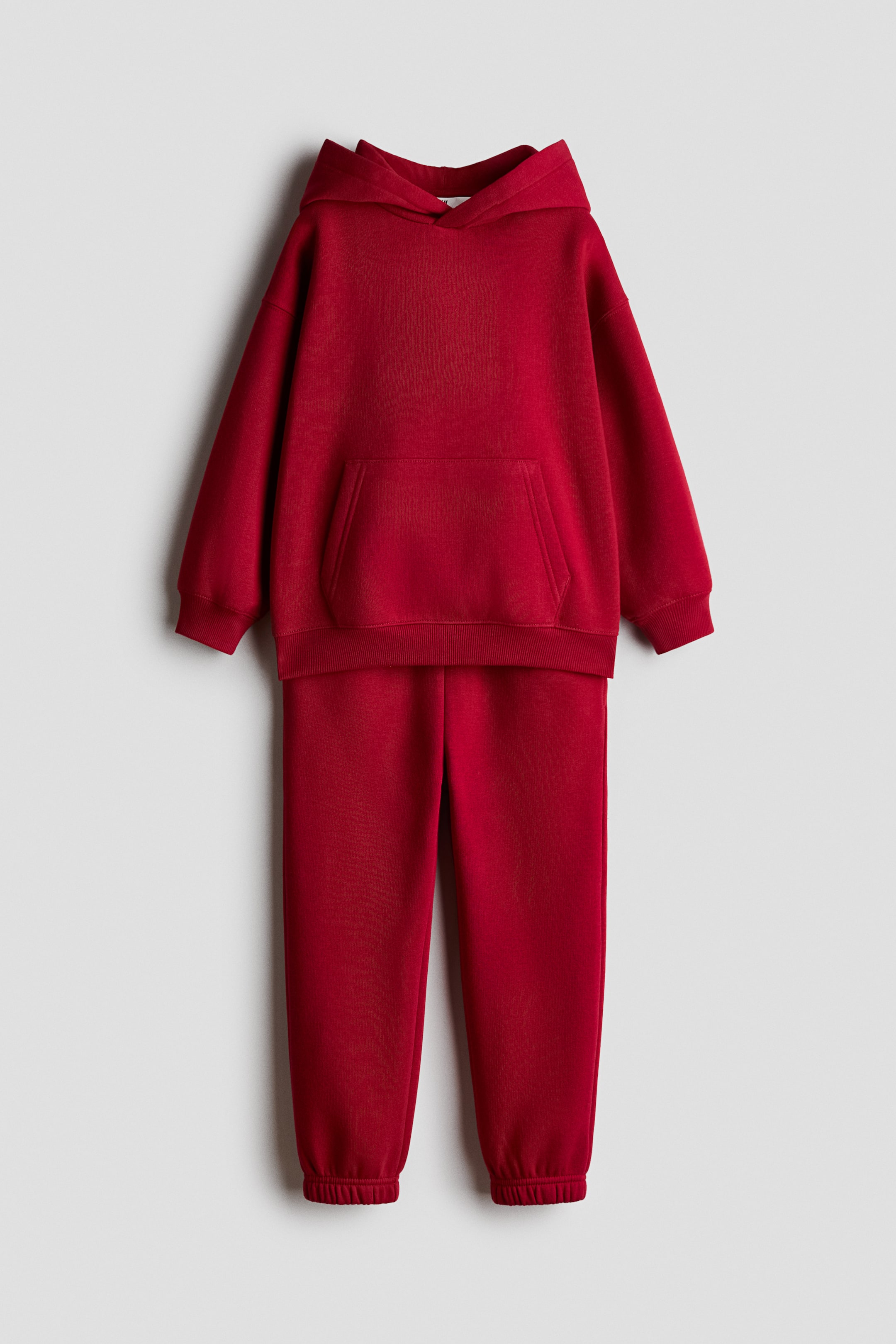 Ampliar la imagen: Se presenta de frente un conjunto de pants rojo, compuesto por una sudadera tipo hoodie y pants a juego. La hoodie cuenta con un bolsillo frontal tipo canguro, mangas largas con puños acanalados y dobladillo acanalado, mientras que los pants muestran puños elásticos en los tobillos.