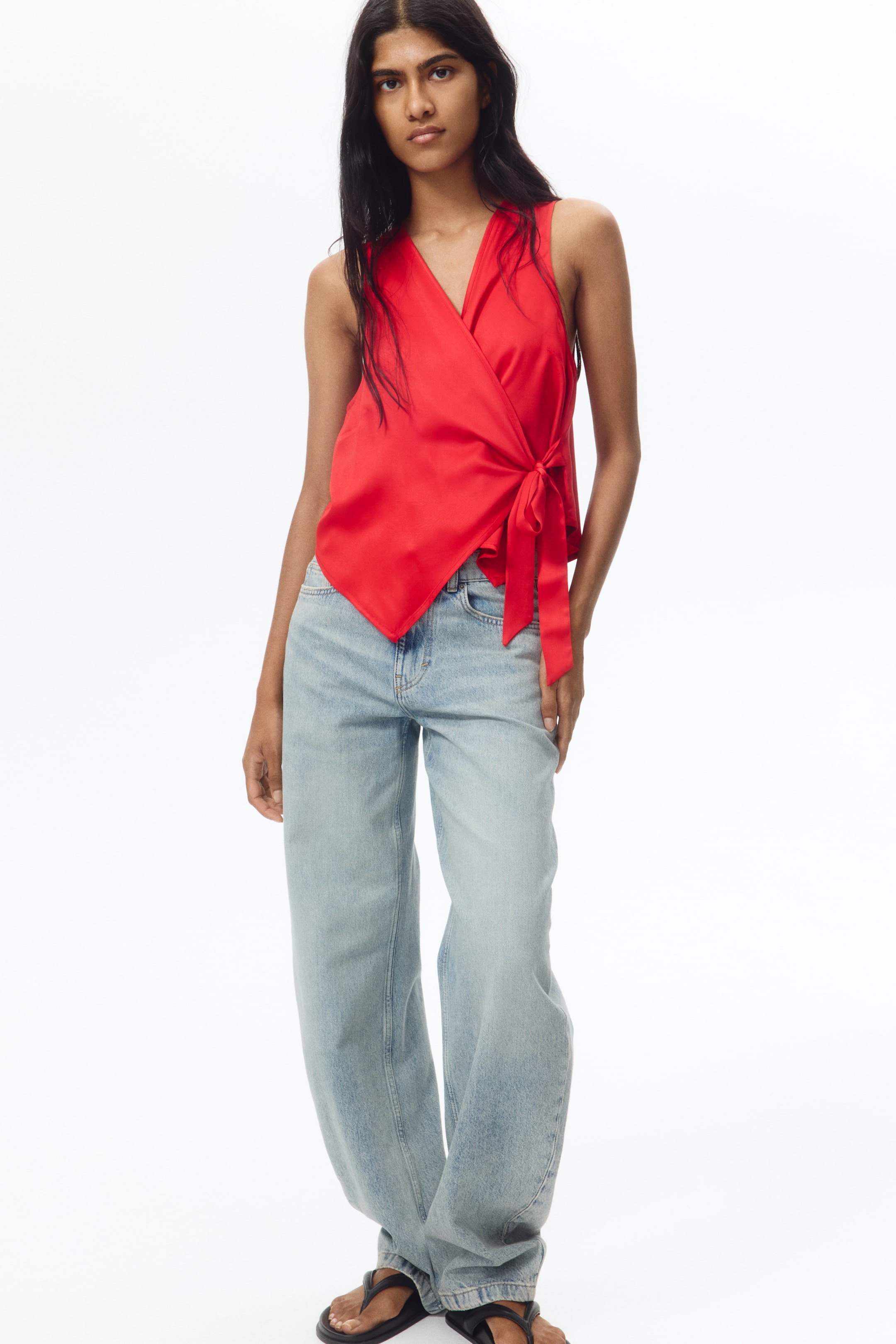 View larger image: A red satin sleeveless wrap top showcasing a deep V-neckline and a front wrap design. It ties at the left side waist, forming a decorative bow and creating an asymmetrical hemline.
