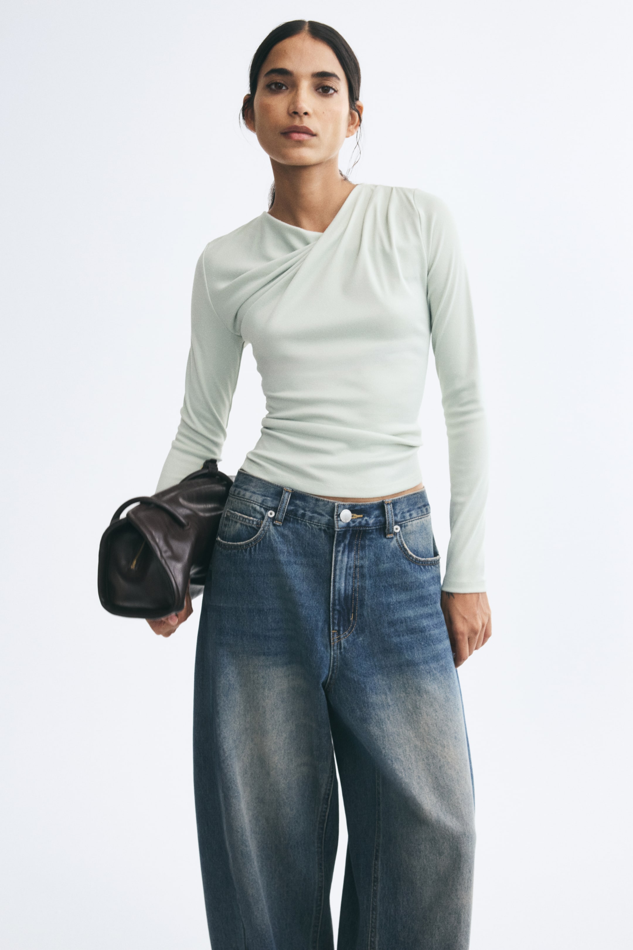 View larger image: The model is wearing a light green fitted long-sleeve top featuring an asymmetrical neckline and draped pleating on one shoulder. The top is styled with wide-leg blue jeans, and the model is carrying a dark brown leather bag under one arm.