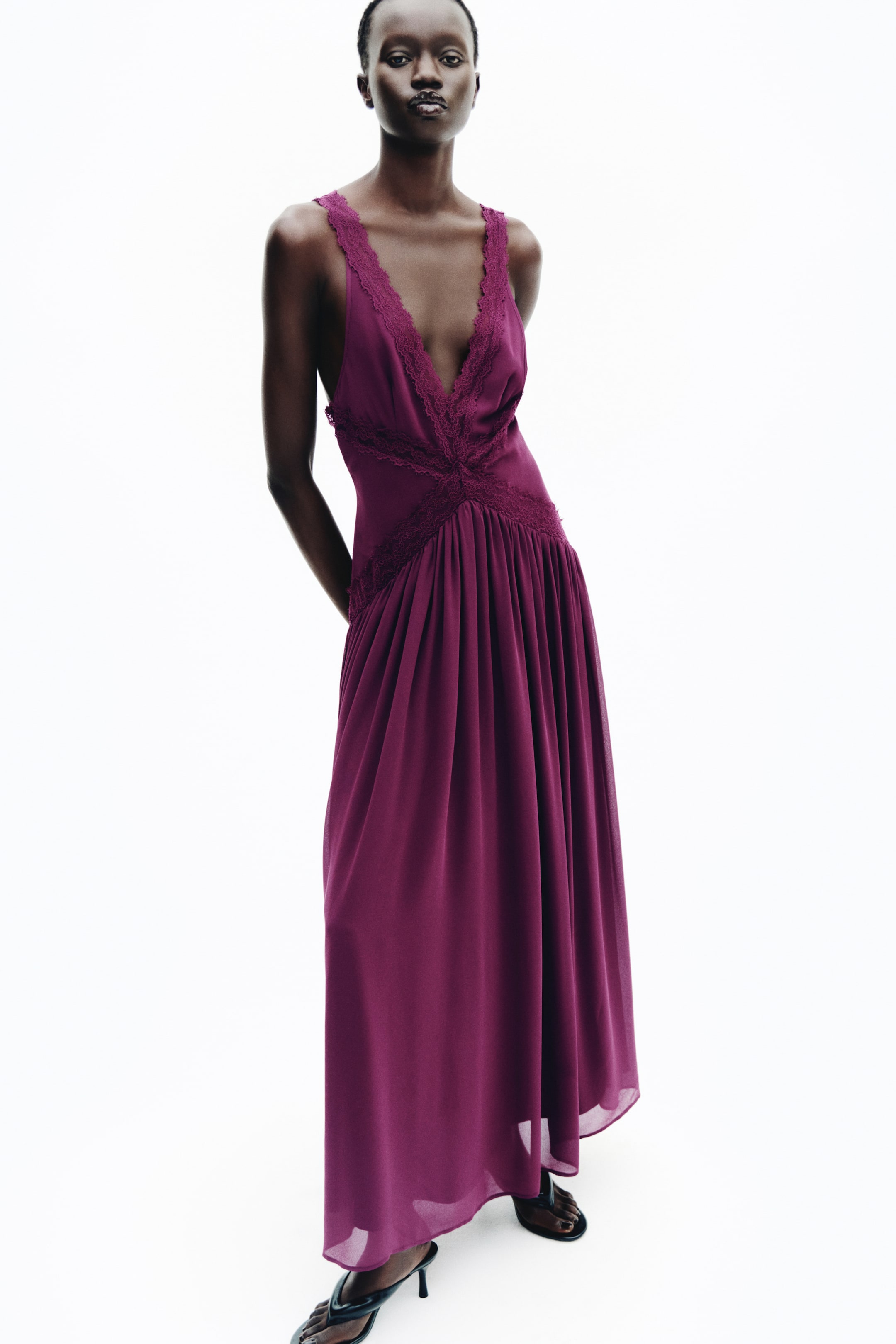 View larger image: A plum-colored chiffon maxi dress with a deep V-neckline, lace trim, and lace inserts at the waist is worn by the model. The dress features a gathered, flowing skirt and is styled with black heeled sandals.