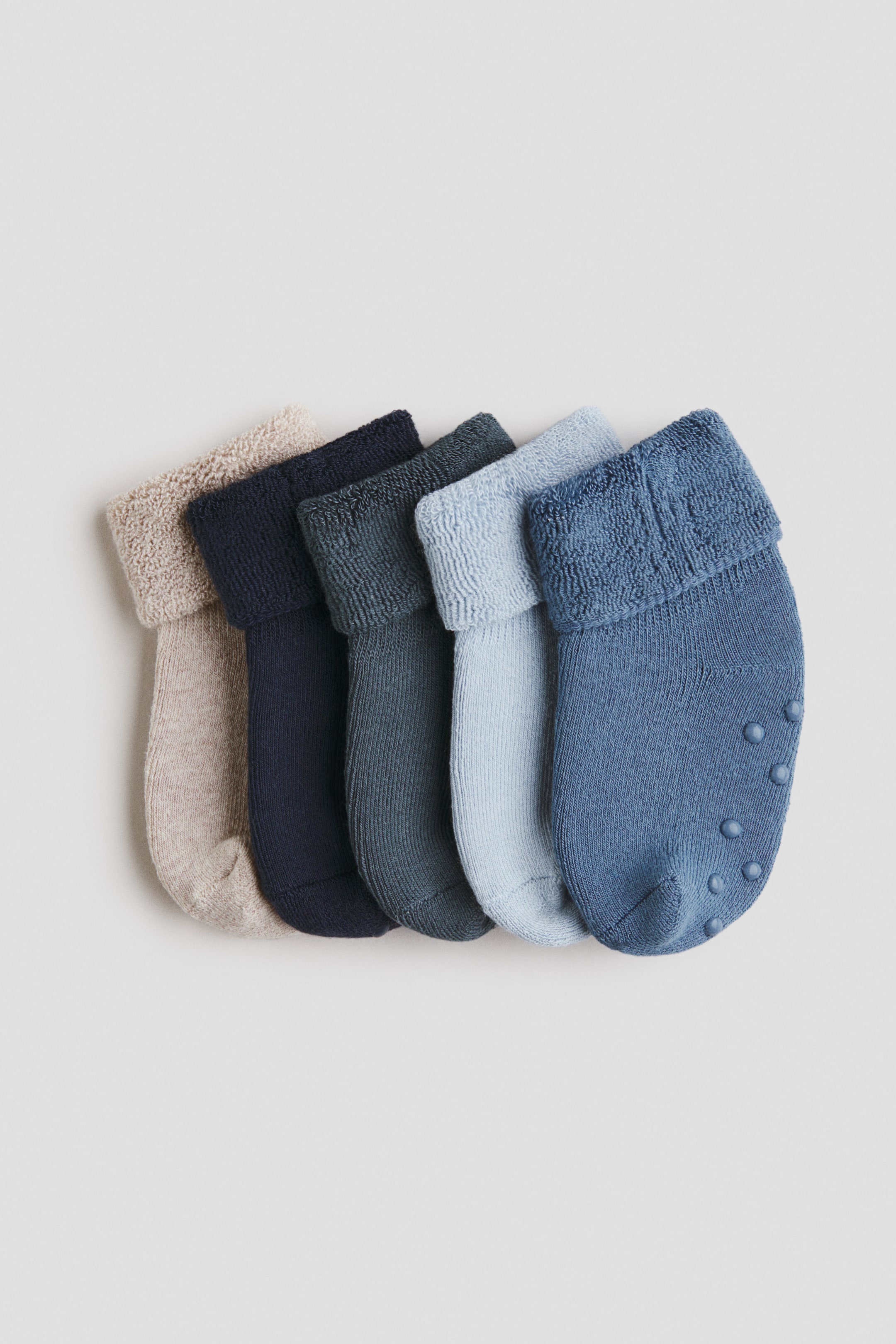 View larger image: A set of five fine-knit socks is laid flat, arranged in a colour gradient from beige to various shades of blue. Each sock features a folded-down shaft with a visible soft, textured inner surface, and the rightmost blue sock displays anti-slip protectors on its sole.