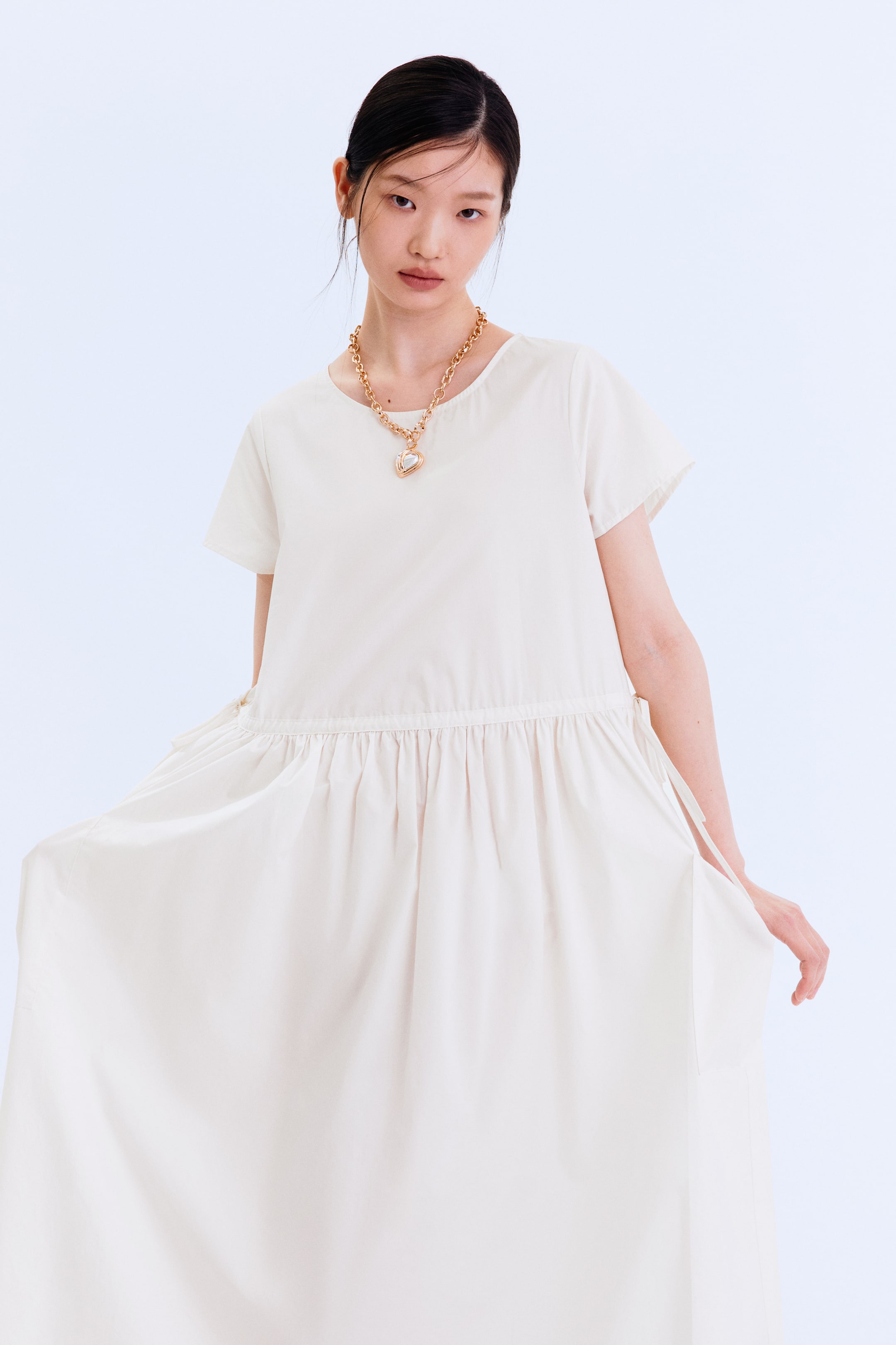 拡大画像を表示: オフホワイトの正面向きのドレスは、ショートスリーブ、丸いネックライン、ギャザーの入ったドローストリングウエストが特徴で、そこから広がるフルで流れるようなスカートがボリュームを見せるために少し外側に広げられています。ドレスの上には、ハートロケット付きのゴールドトーンのチェーンネックレスが着用されています。