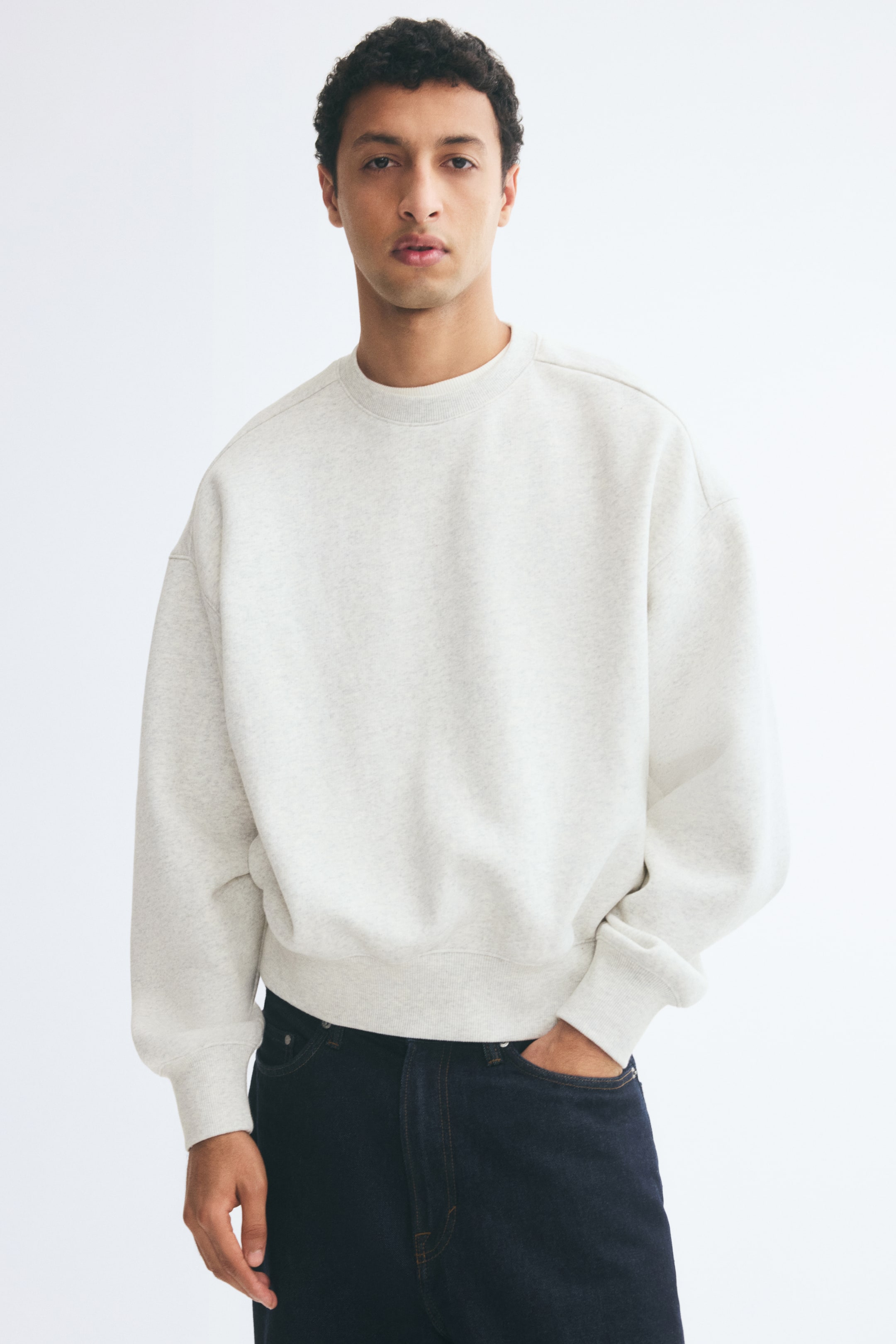 View larger image: The model is wearing a light gray oversized sweatshirt with a round, rib-trimmed neckline, dropped shoulders, and ribbing at the cuffs and hem. It is paired with dark jeans, with one hand casually tucked into a front pocket.