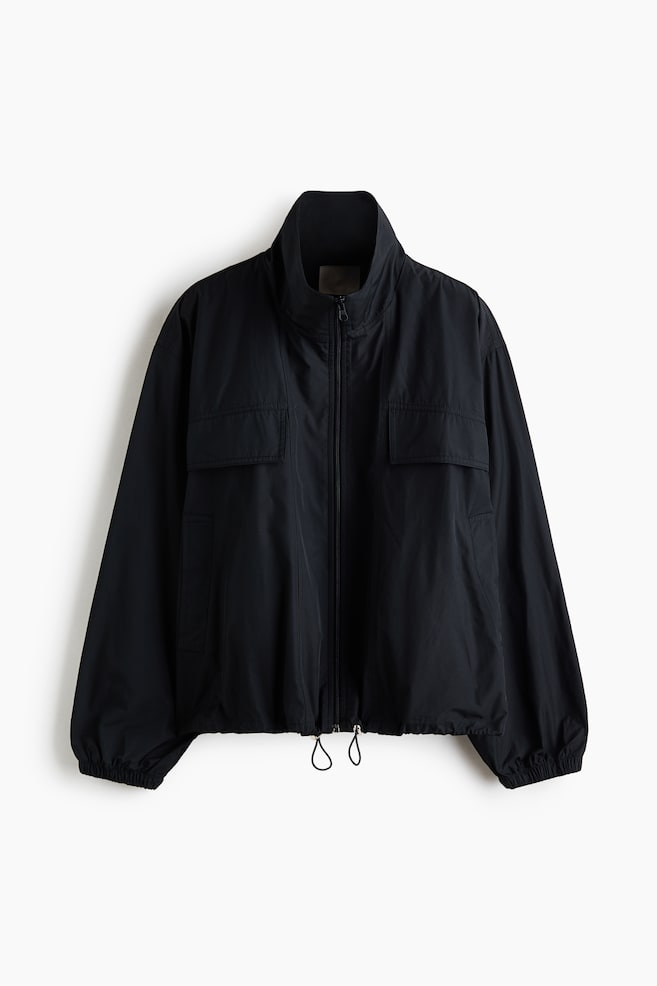 A black lightweight jacket is facing front, showcasing a high stand-up collar, a full-length front zip, and two chest pockets with rectangular flaps. The garment features relaxed-fit long sleeves with elasticated cuffs and an adjustable hem with hanging drawstrings. - 1
