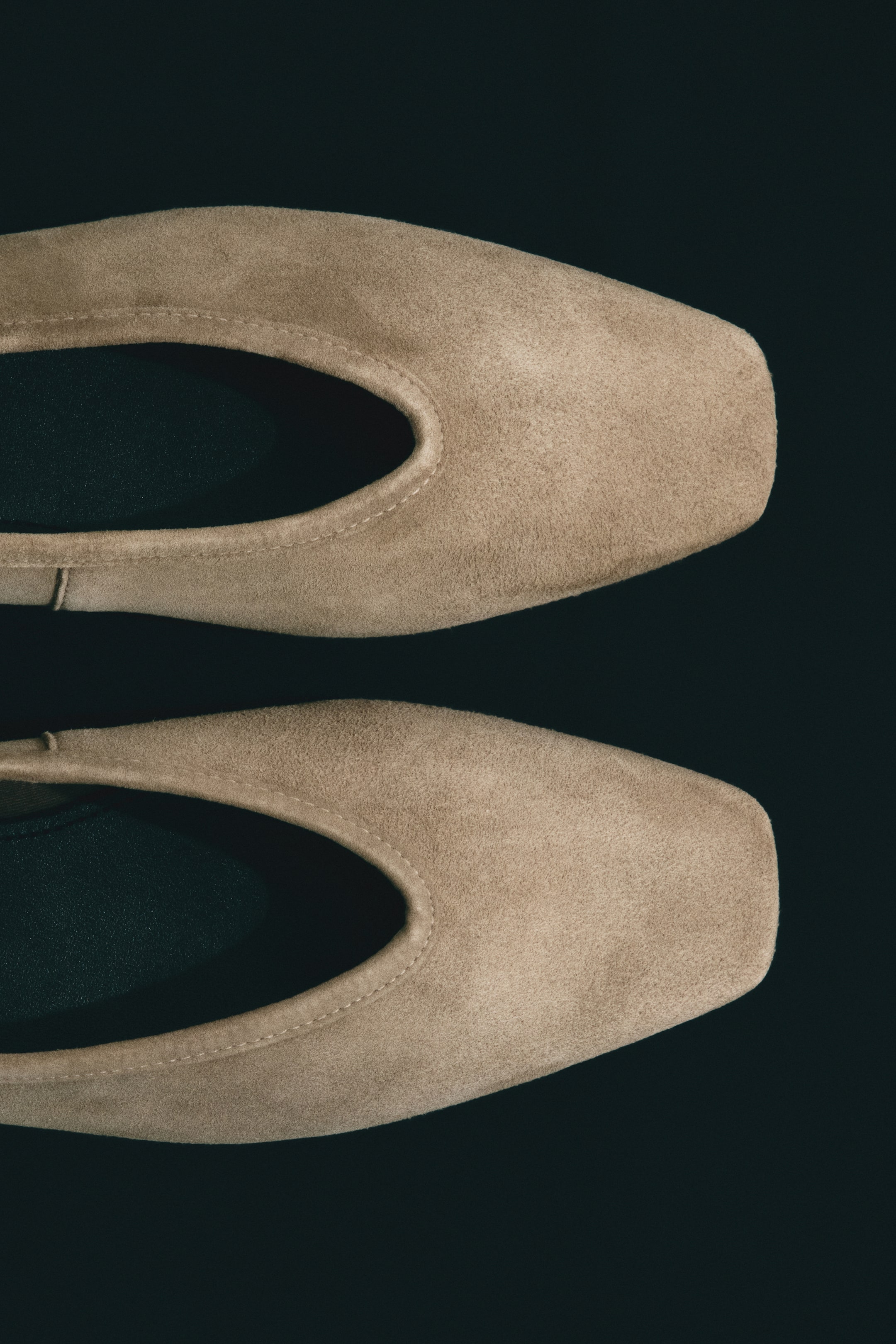 Ver imagem maior: "Duas sabrinas em camurça castanho claro estão voltadas para a frente numa perspetiva de cima para baixo, exibindo as suas pontas ligeiramente quadradas e aberturas em forma de U. Costuras visíveis delineiam as aberturas, revelando um forro escuro no interior de cada sapato."