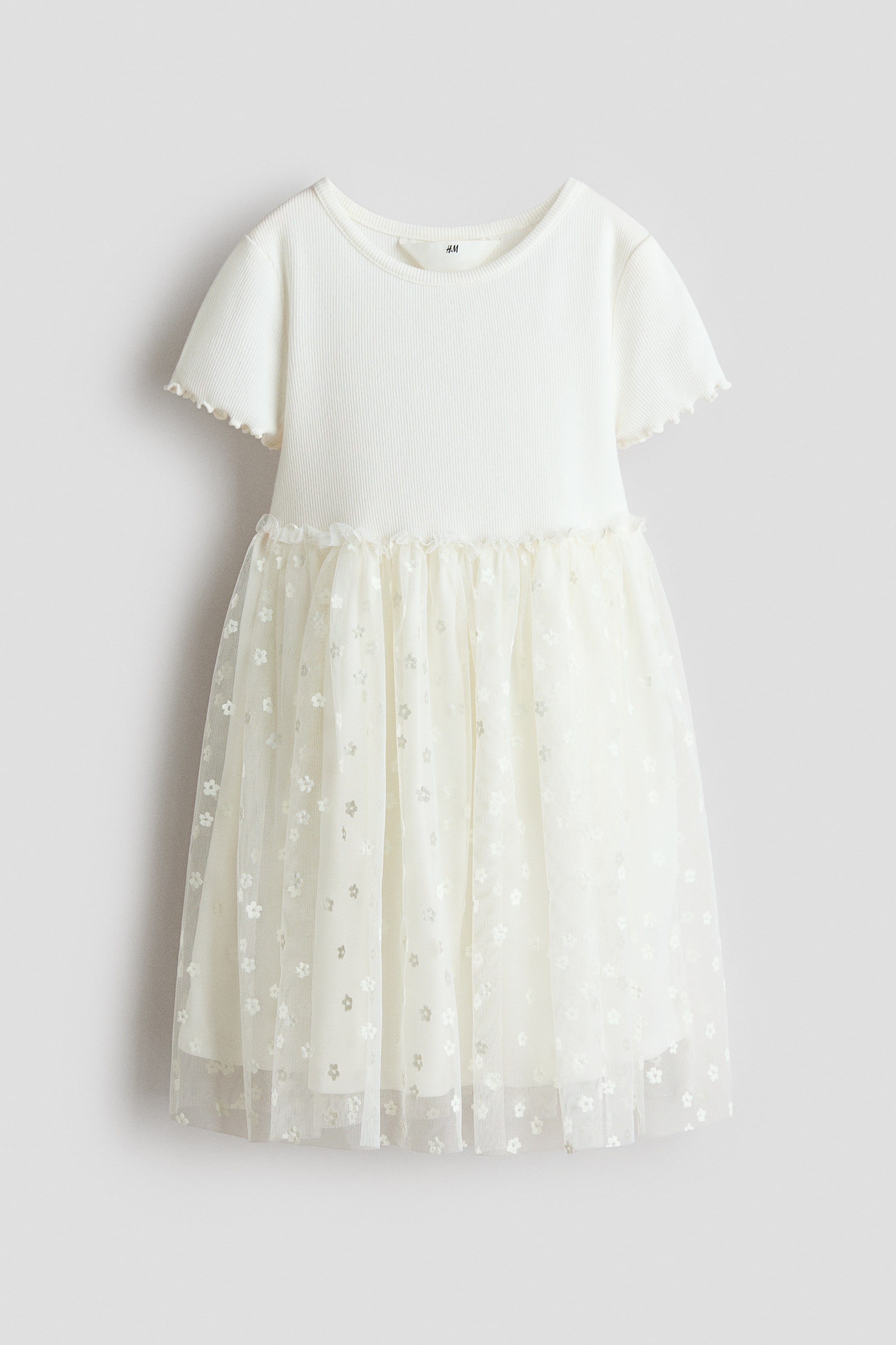 View larger image: The image displays a short-sleeved dress from the FRONT, featuring a white, ribbed jersey bodice with a round neckline and picot trim, along with frill-trimmed short sleeves. A gathered seam with a frill accentuates the waist, leading to a glittery tulle skirt adorned with a floral pattern, layered over a solid lining.