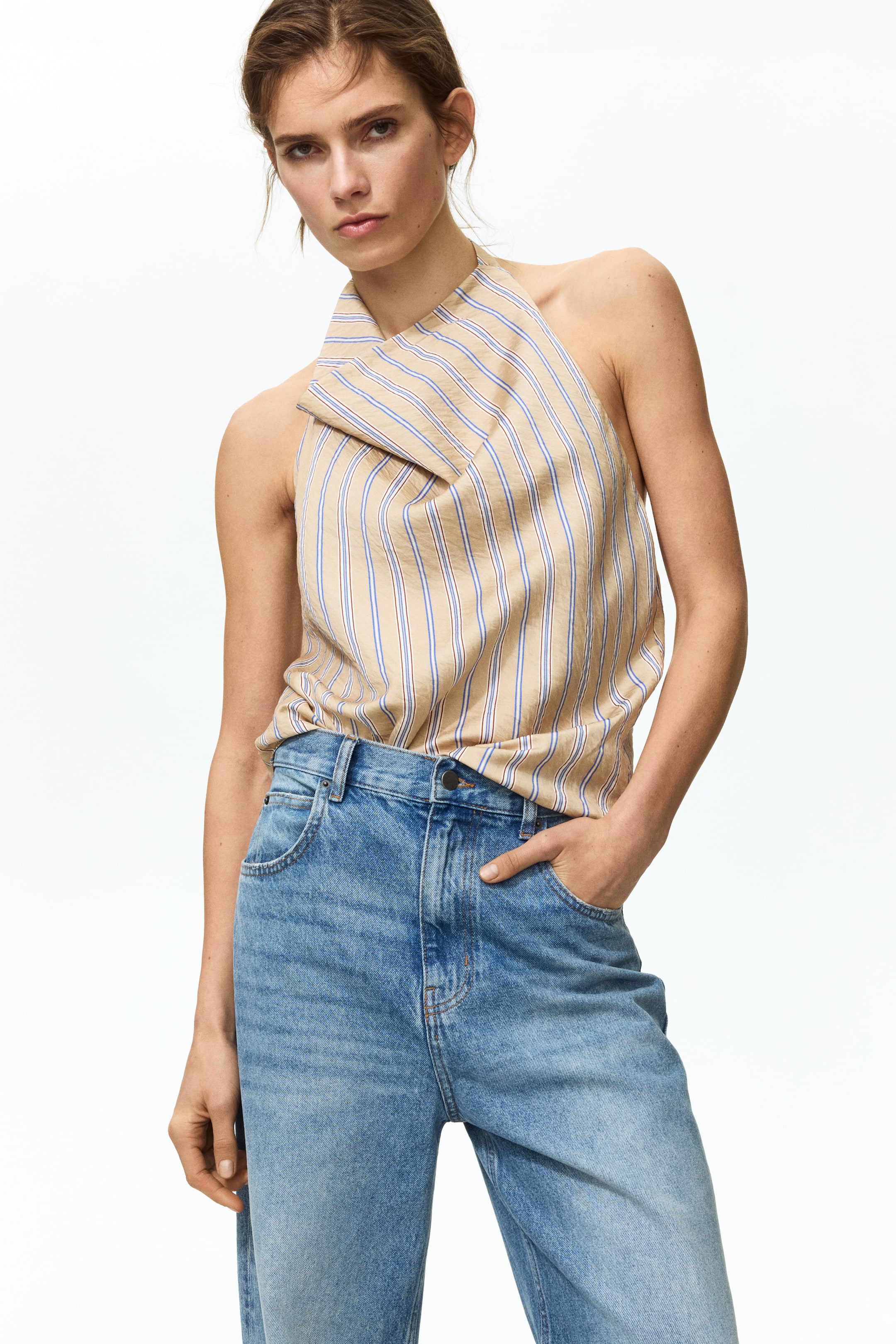 View larger image: Presenting a beige halter neck top with thin blue and red vertical stripes and an asymmetric, folded neckline. The top is tucked into light blue, high-waisted jeans featuring front pockets and a faded wash.