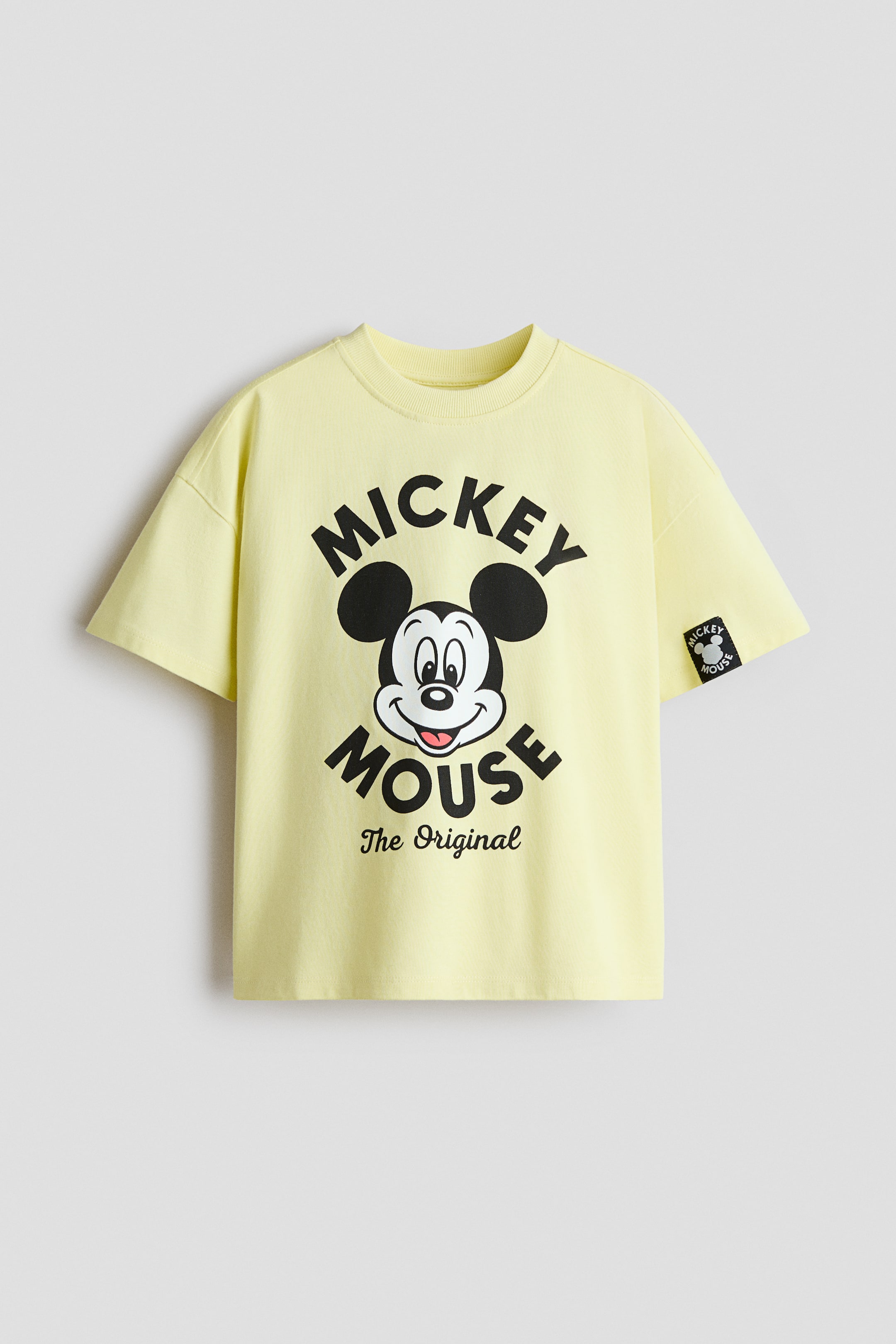 Größeres Bild ansehen: Ein hellgelbes, locker sitzendes T-Shirt aus weichem Baumwolljersey wird flach und frontseitig präsentiert. Es zeigt einen großen schwarzen Mickey Mouse Grafikdruck auf der Brust, einen runden, gerippten Halsausschnitt und überschnittene Schultern, mit einem kleinen schwarzen Mickey Mouse Aufnäher auf dem rechten Ärmel.