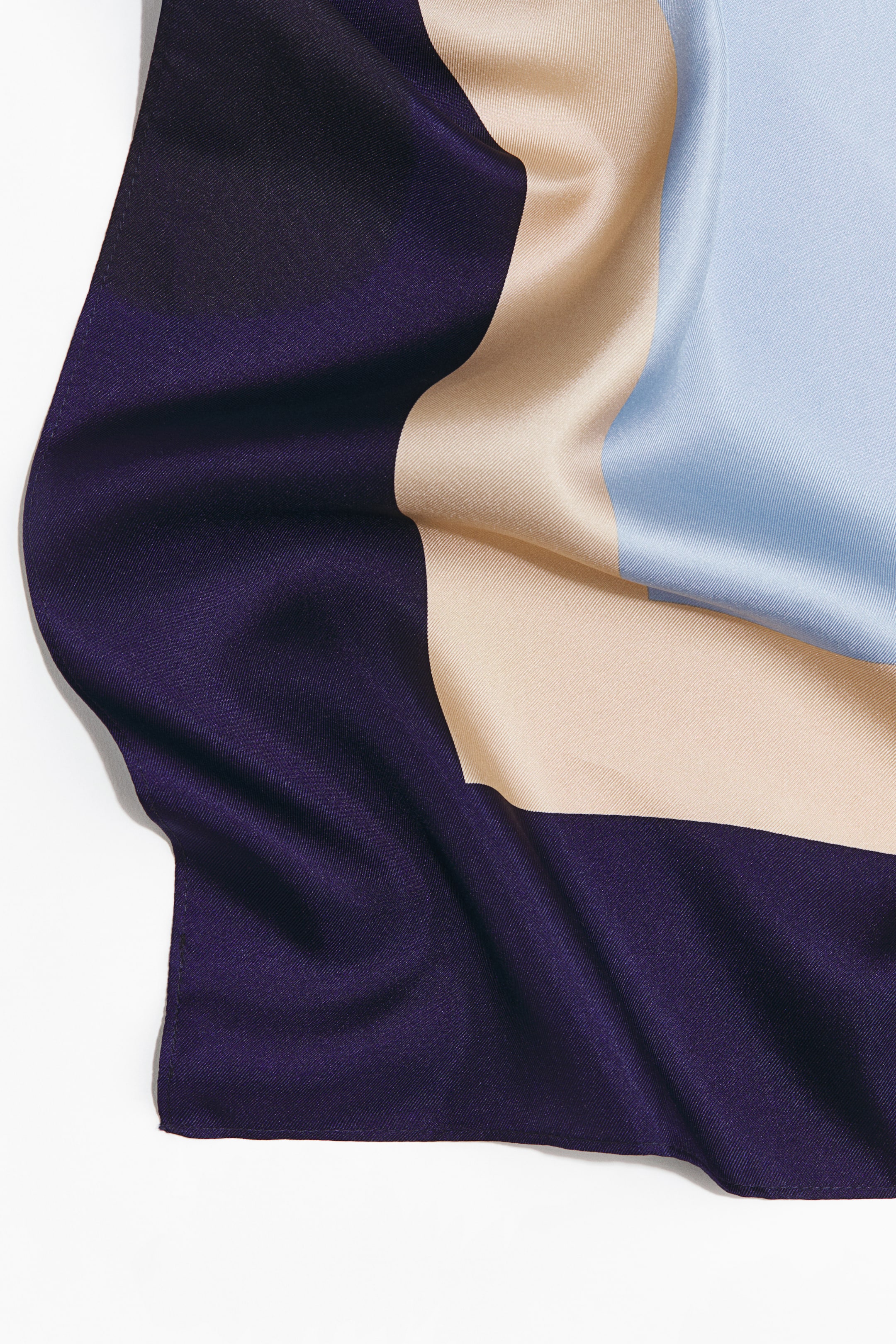 Ampliar la imagen: Un pañuelo cuadrado de suave seda de morera muestra un diseño gráfico de grandes bloques de color contrastantes en morado oscuro, beige claro y azul claro. La tela muestra pliegues suaves, con una sutil costura oscura visible a lo largo de sus bordes.