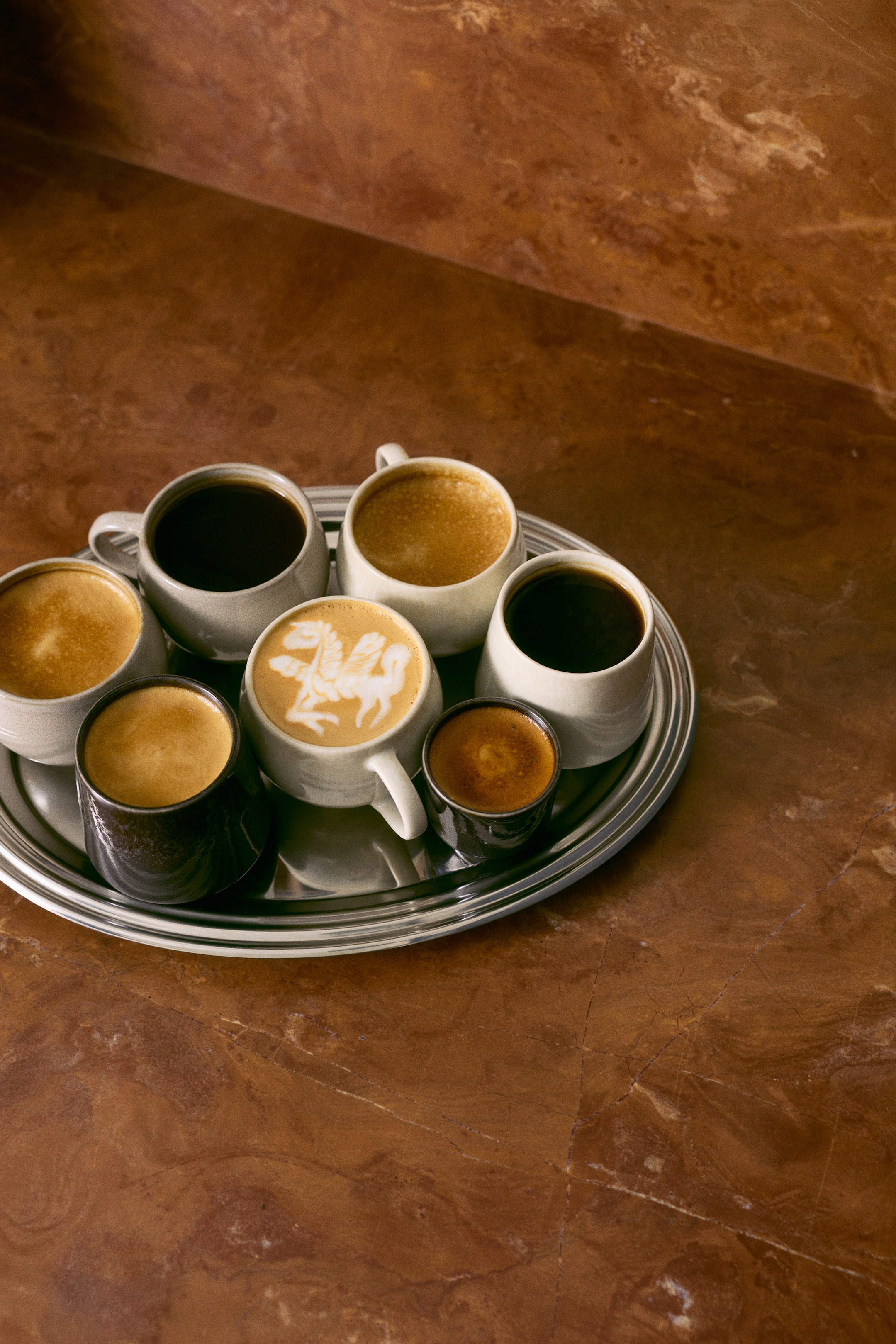 View larger image: Stoneware mugs and cups featuring a glossy, reactive glaze in speckled off-white and dark charcoal shades are resting on a silver-toned metal tray. The organic-shaped vessels are shown from a high angle, filled with various coffee beverages, including espresso and a latte with latte art.