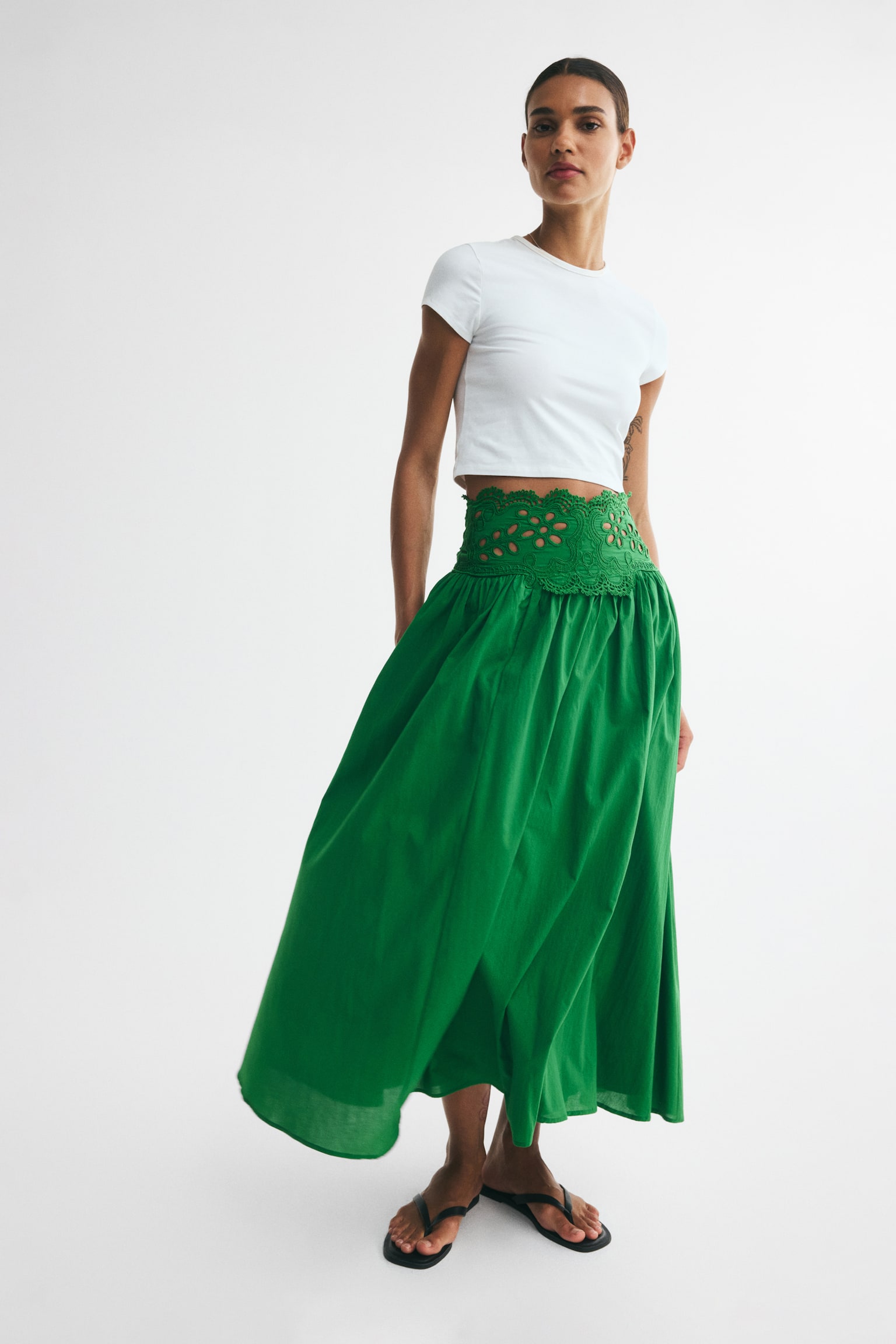 A long green skirt, displayed from the front, boasts a broad lace section with intricate cutouts at its high waist. The skirt gathers at the hips, adding volume, and gracefully flows downwards in a cotton weave. - 1