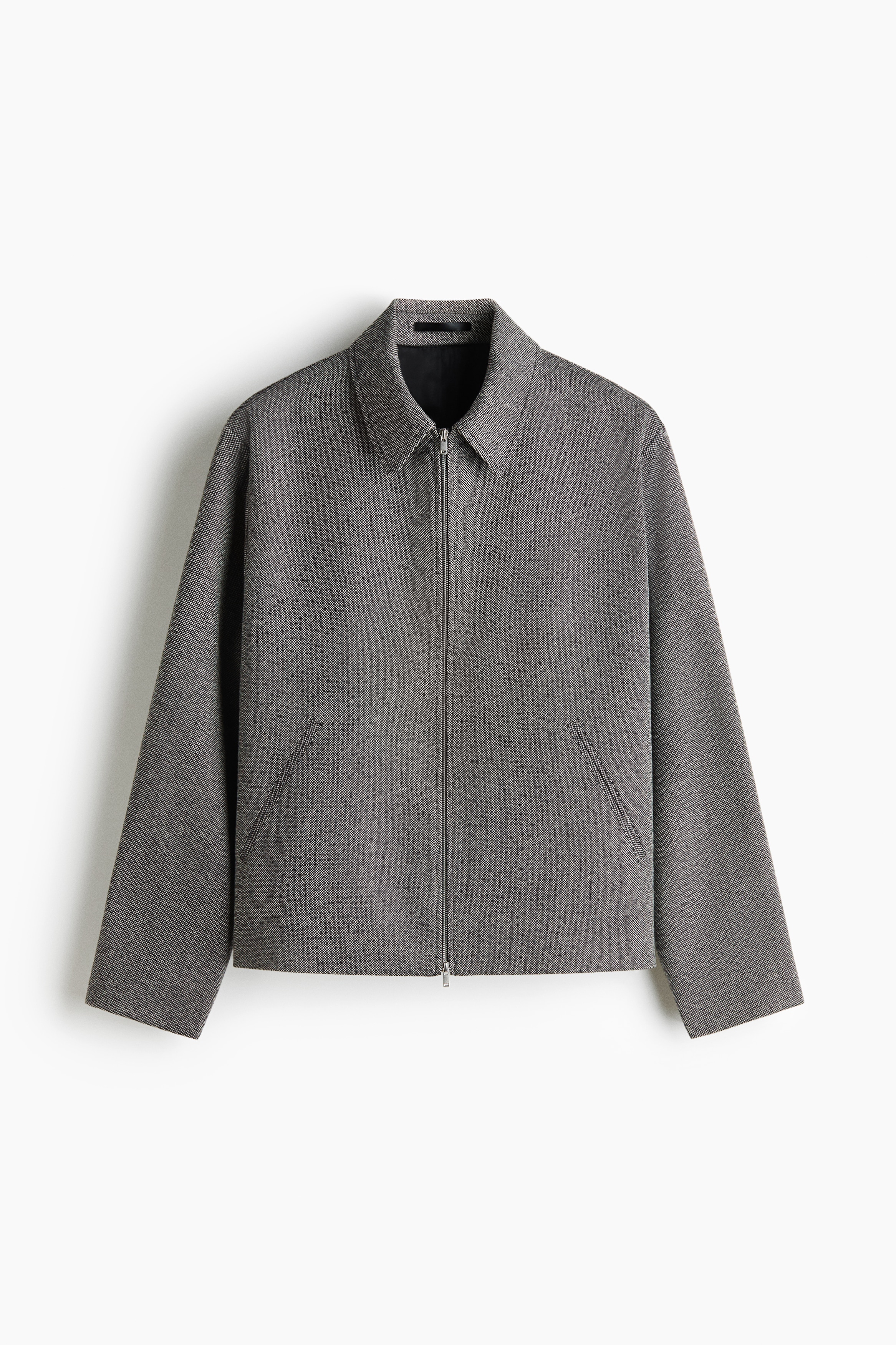 A grey, textured wool-blend jacket featuring a pointed collar and a silver two-way zip is being worn by the model. The jacket is layered over a black turtleneck jumper and styled with matching grey trousers.