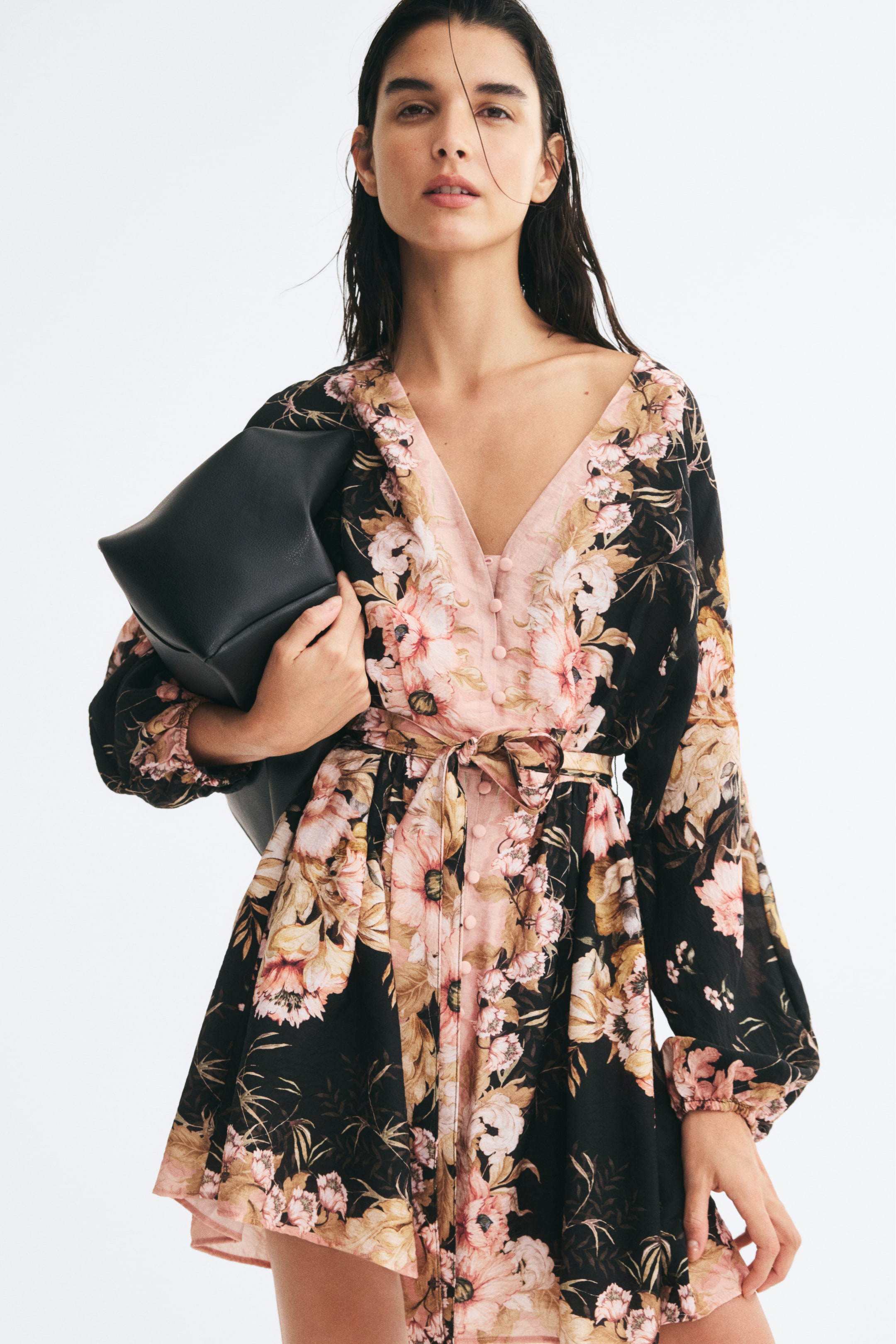 View larger image: A floral-print dress in black, pink, and beige is being worn by the model, featuring a V-neckline, front button-and-loop fasteners, long balloon sleeves, and a matching tie belt at the waist. The model is holding a black bag under one arm while wearing the dress.