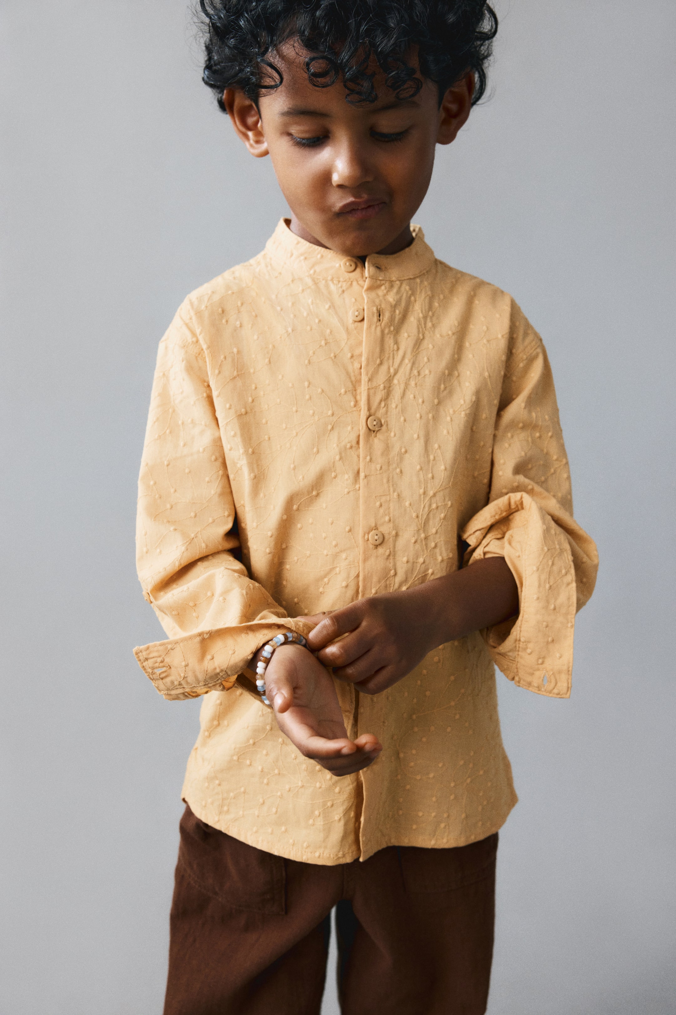 View larger image: A front view shows a light apricot-coloured grandad shirt in a soft, patterned cotton weave, featuring a band collar and a button-down front. The long sleeves with button cuffs are being rolled up, and the shirt is finished with a gently rounded hem.