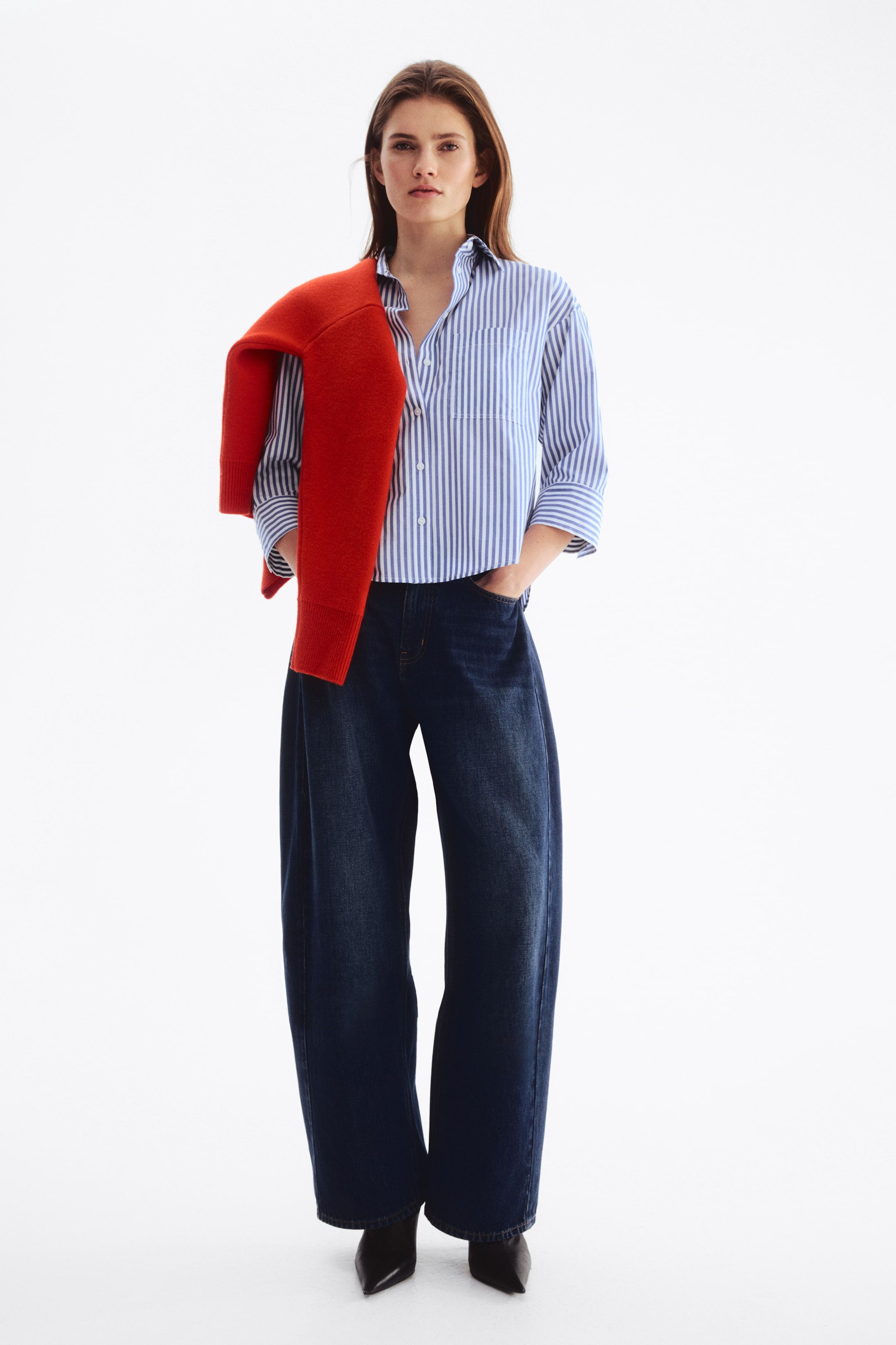 View larger image: A blue and white striped button-up shirt, featuring a collar, a chest pocket, and rolled-up sleeves, is being paired with dark wash, wide-leg jeans and black pointed-toe shoes. A red sweater is being draped over the shoulders, resting on the shirt.