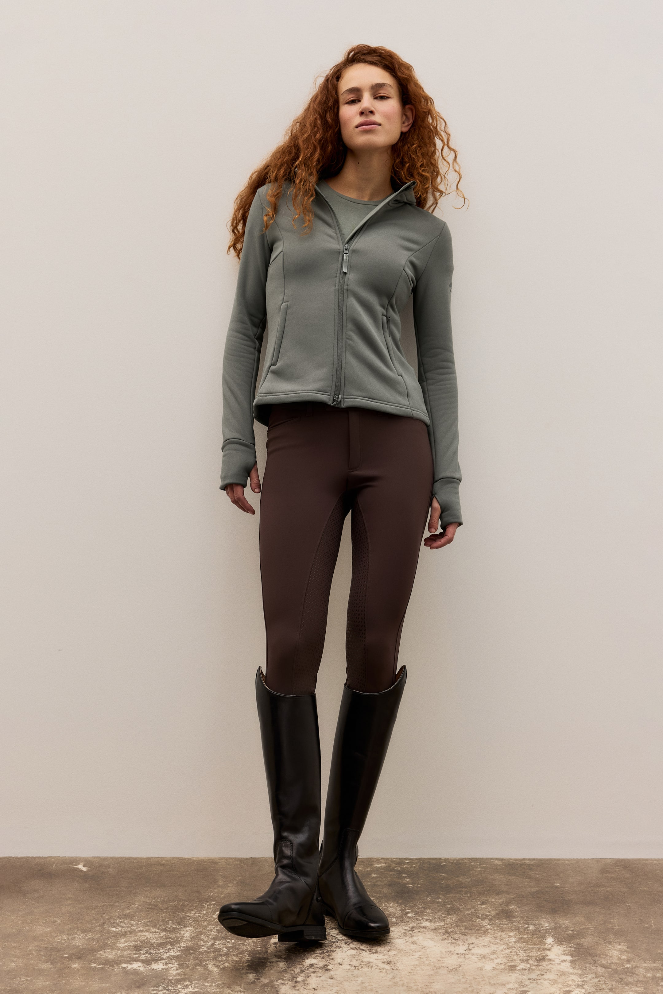 Agrandir l'image: Une tenue équestre complète est présentée, composée d'une veste à capuche zippée complète vert sauge avec poches avant zippées et poignets à trous pour les pouces, de culottes d'équitation marron foncé avec panneaux intérieurs texturés et de bottes d'équitation hautes en cuir noir.