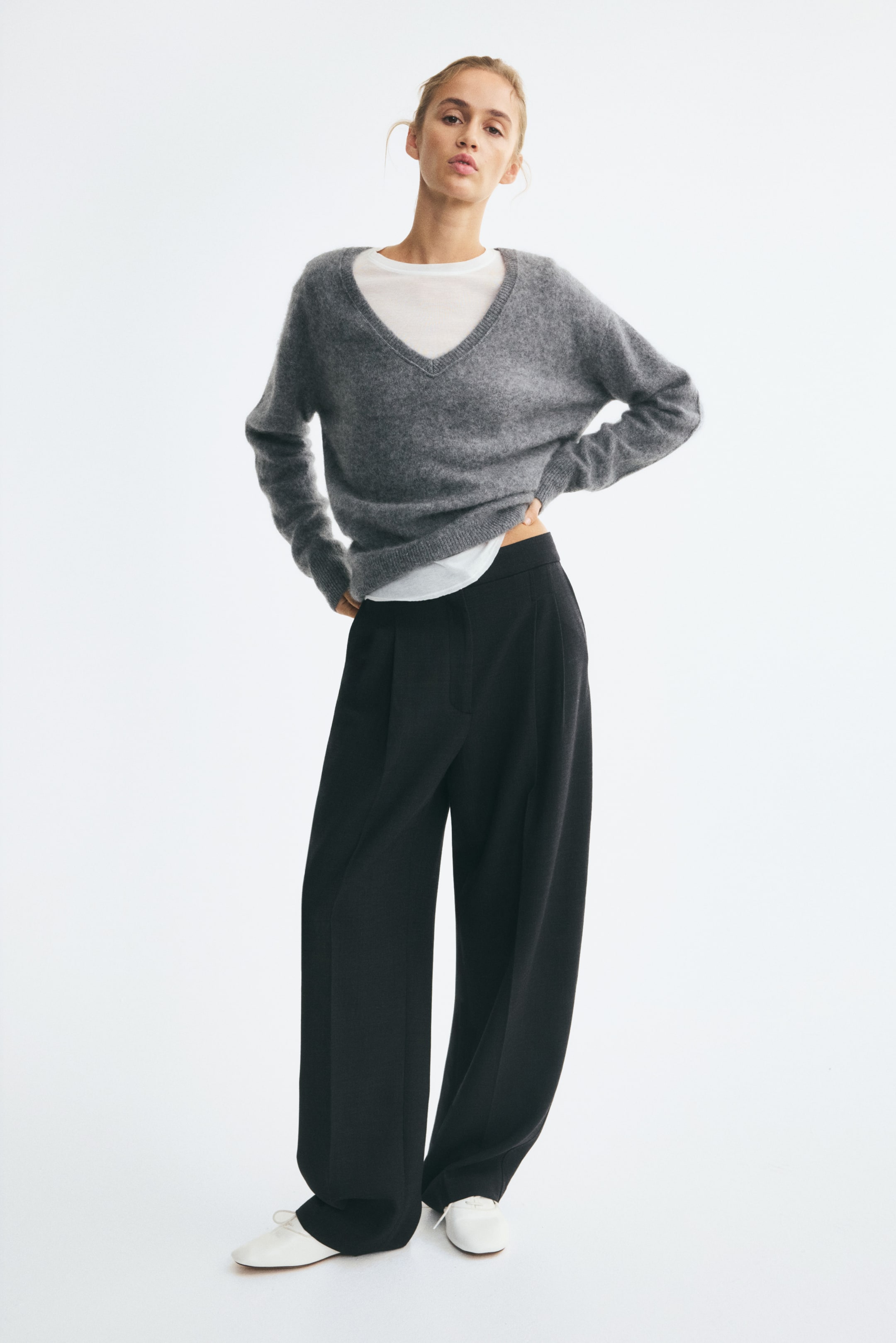 Ampliar la imagen: Pantalones negros de corte holgado en tejido de mezcla de lana que lleva la modelo, con cintura alta, pinzas delanteras y perneras tipo barrel con pliegues visibles al frente. Los pantalones se combinan con un suéter gris de cuello en V sobre una camiseta blanca y zapatos blancos.