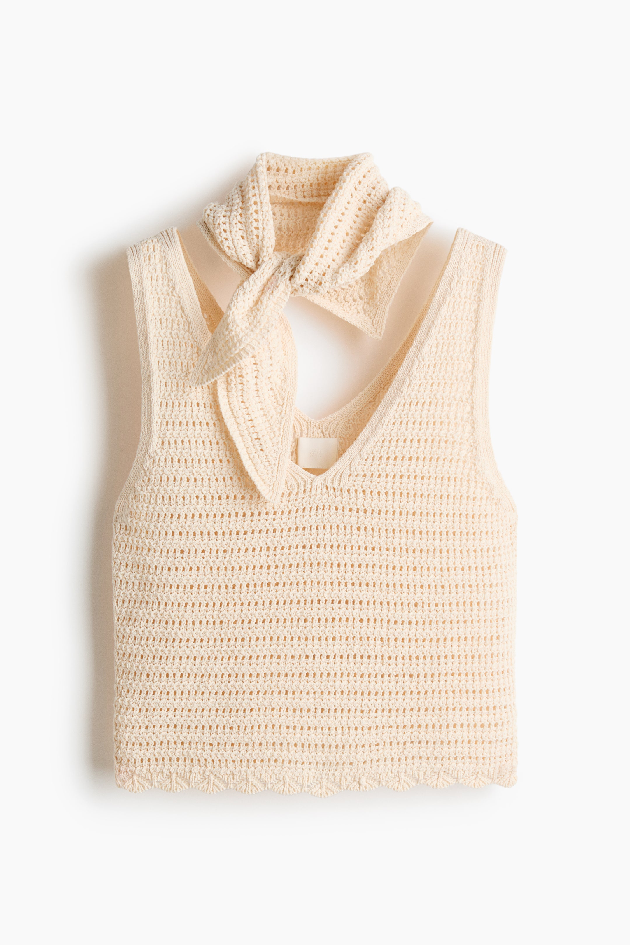Ampliar la imagen: Un top sin mangas de crochet en color crema se presenta en una disposición plana, con un escote en V y una pieza de crochet a juego atada al cuello. El top exhibe un patrón de crochet abierto a lo largo de su cuerpo y un dobladillo festoneado.