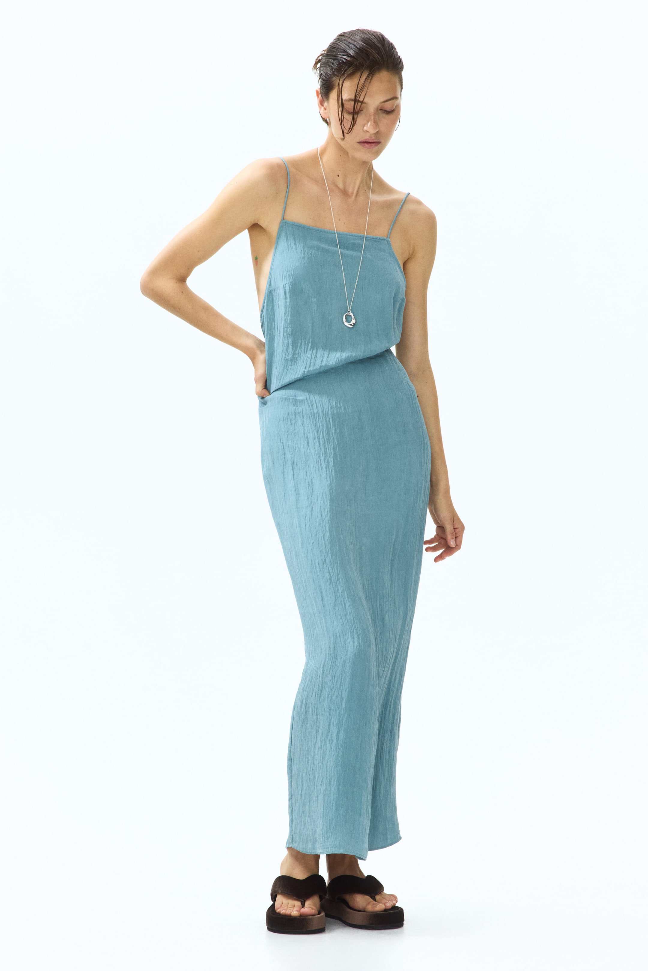 View larger image: The product is a long, light blue dress, showcasing its front and left side. It features spaghetti straps, a straight neckline, and a blousing top that transitions into a fitted skirt with a crinkled texture.