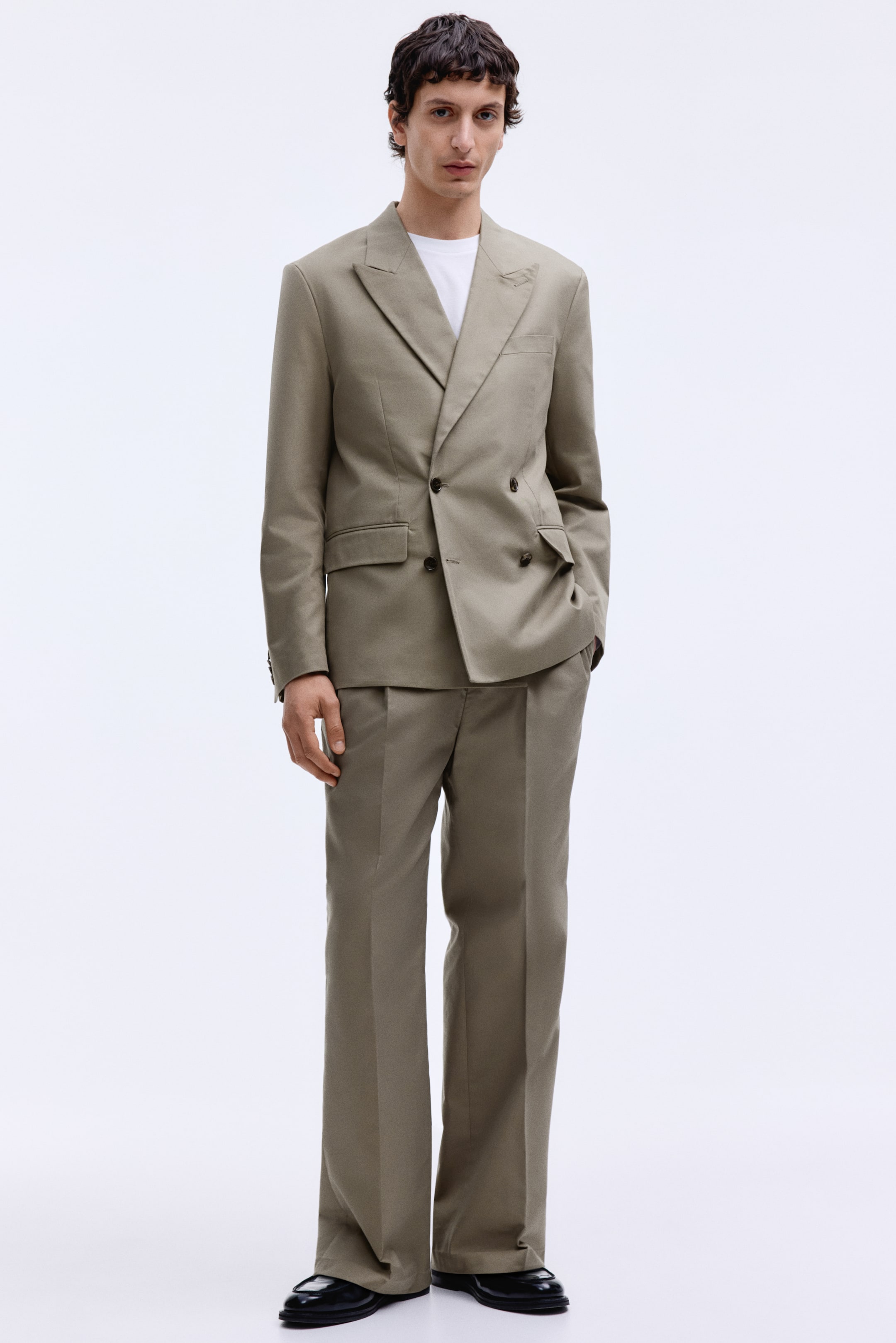 View larger image: A khaki double-breasted suit is showcased from a front-facing angle, featuring a jacket with peak lapels, four buttons, and flap pockets. The jacket is paired with a white crew-neck t-shirt, and the coordinating wide-leg trousers, with visible pleats at the waist, complete the suit, styled with black shoes.