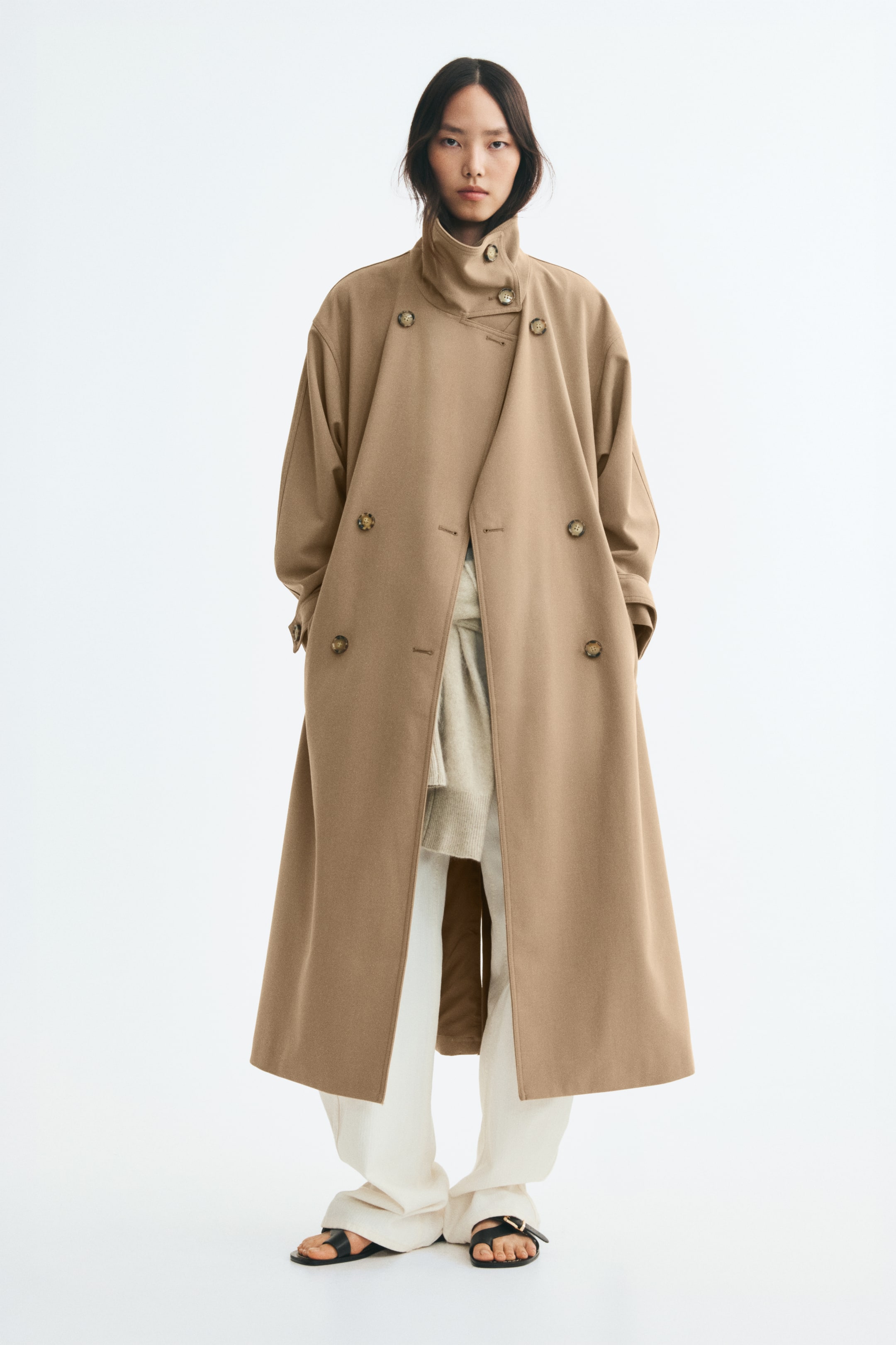 Näytä suurempi kuva: Mallilla on yllään väljä, kaksirivinen beige twill-trenssitakki, jossa on korkea, napitettava suppilokaulus ja pudotetut olkapäät. Avoin takki on kerrostettu luonnonvalkoisten housujen ja neuleen päälle, ja malli pitää käsiään sivuilla olevissa reunataskuissa.
