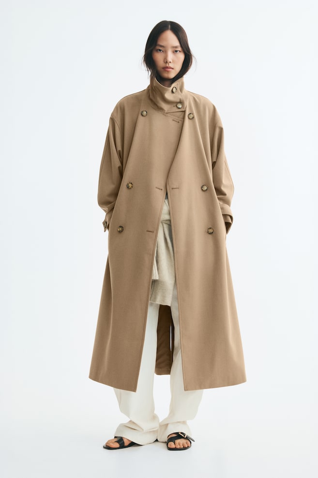 An oversized, double-breasted beige twill trench coat with a high, buttoned funnel collar and dropped shoulders is being worn by the model. The open coat is layered over off-white trousers and a knit sweater, with the model resting their hands inside the side welt pockets. - 1