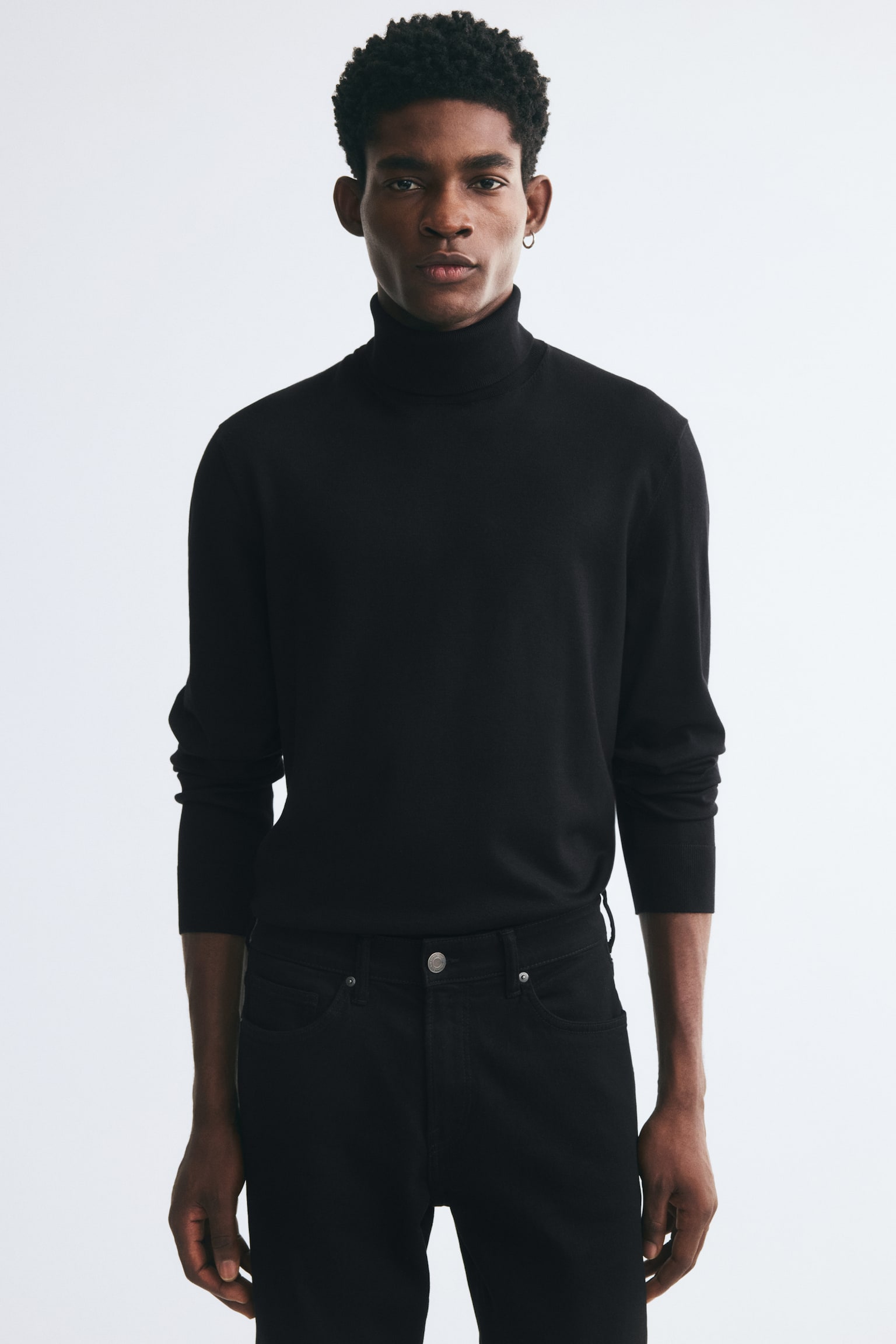 A black fine-knit turtleneck jumper is being worn by the model, featuring a regular fit, a ribbed turtleneck, and long sleeves with ribbed cuffs. The jumper is tucked into a pair of black jeans. - 1