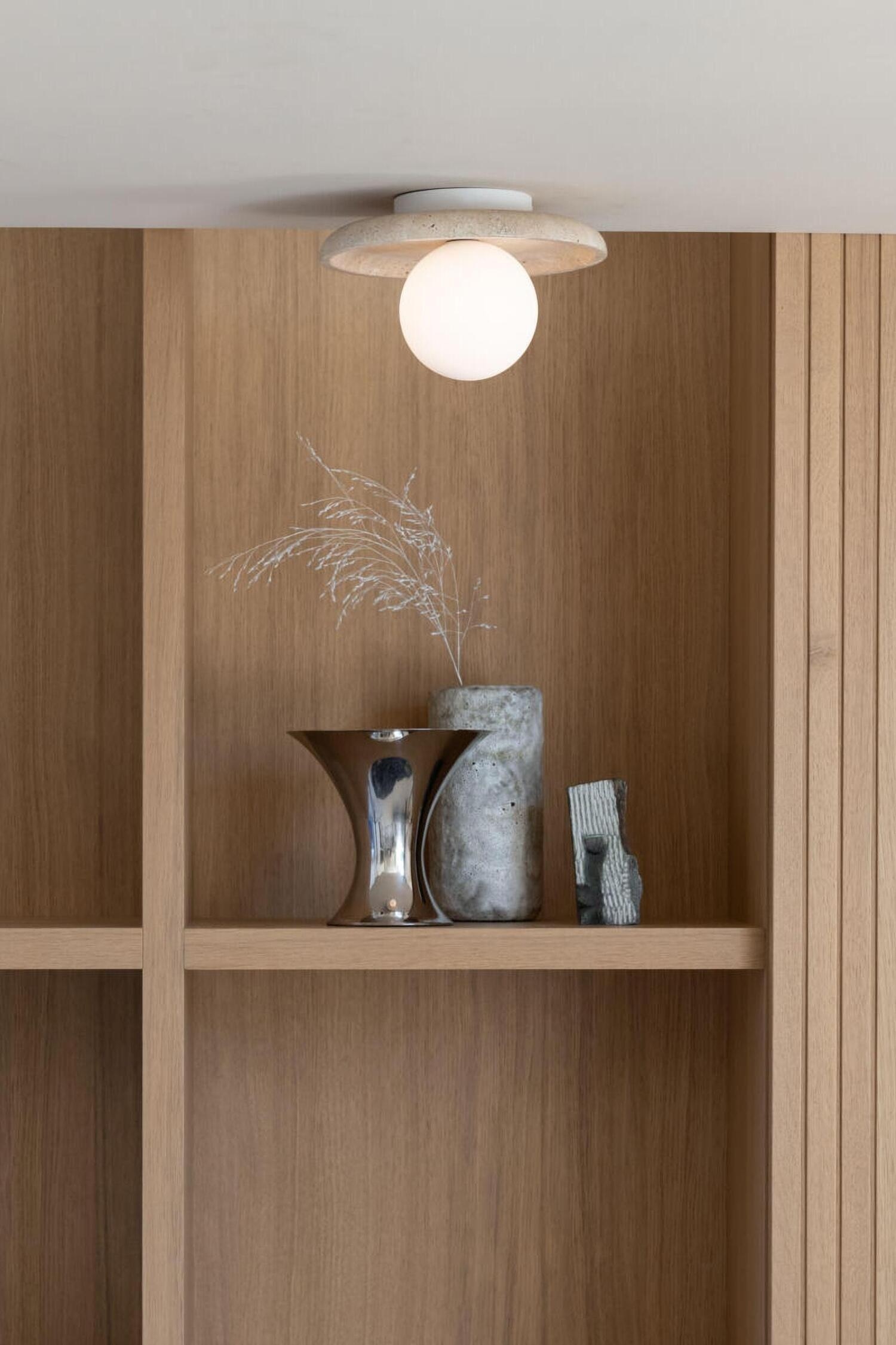 Een opbouwverlichtingsarmatuur met een gloeiende, bolvormige witte glazen globe, gecentreerd onder een brede, ronde plafondplaat van gestructureerde beige travertijnsteen. De verlichte globe werpt warm licht op de onderzijde van de stenen schaal, die verbonden is met een kleine, witte cilindrische basis.
