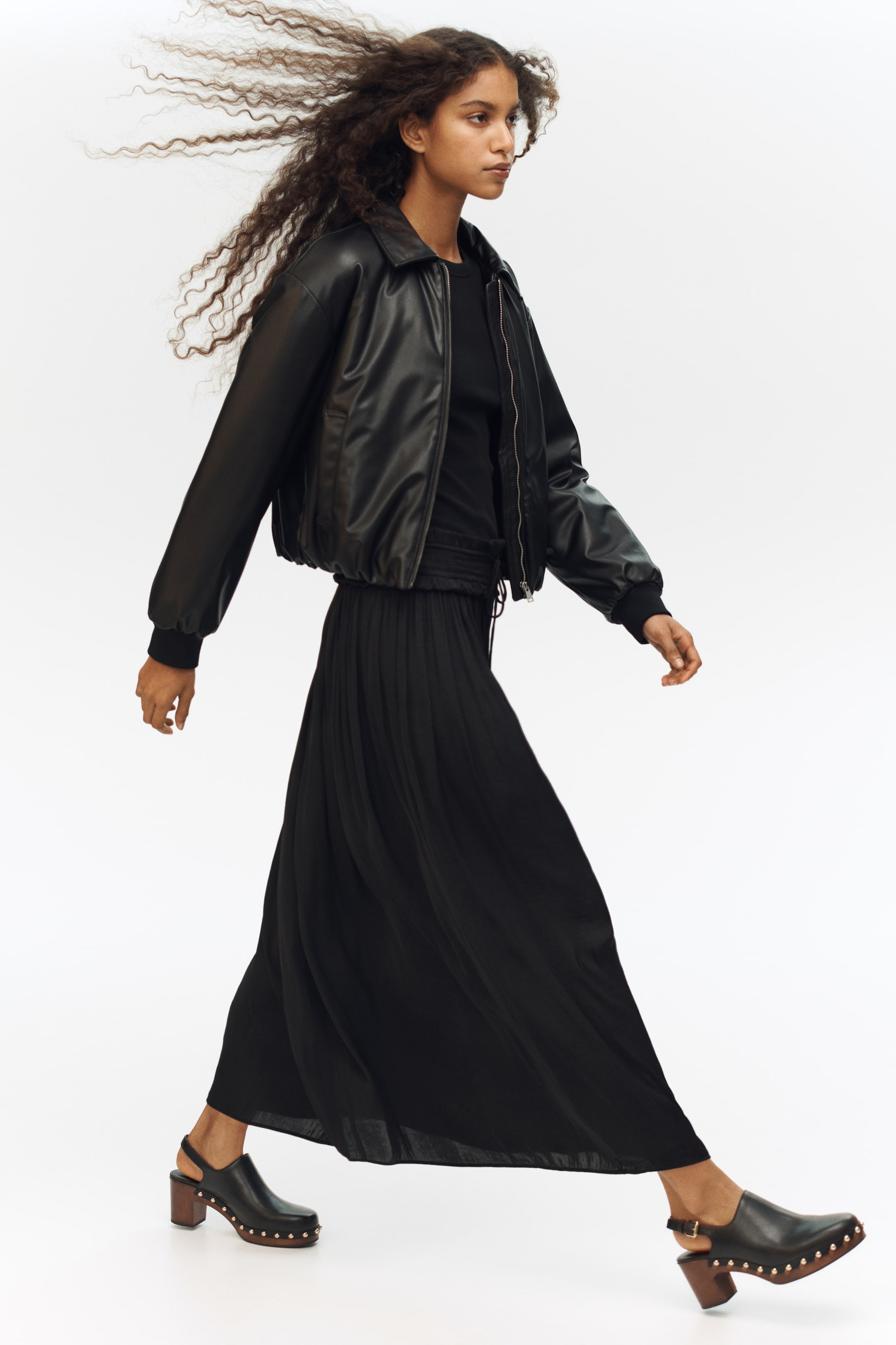 הצגת תמונה גדולה יותר: המוצר מציג אנסמבל שחור מלא, עם דוגמנית הפונה ימינה, ומציגה מעיל בומבר שחור מעור מלאכותי, חצאית מקסי שחורה עם קפלים, וקבקבים שחורים עם ניטים. מעיל הבומבר כולל שרוולים מצולעים ושוליים מצולעים ומכווצים, בעוד שחצאית המקסי עם הקפלים זורמת מחגורת מותן אלסטית, והקבקבים עם רצועה אחורית כוללים סוליית פלטפורמה במראה עץ עם עקב בלוק וניטים מתכתיים.