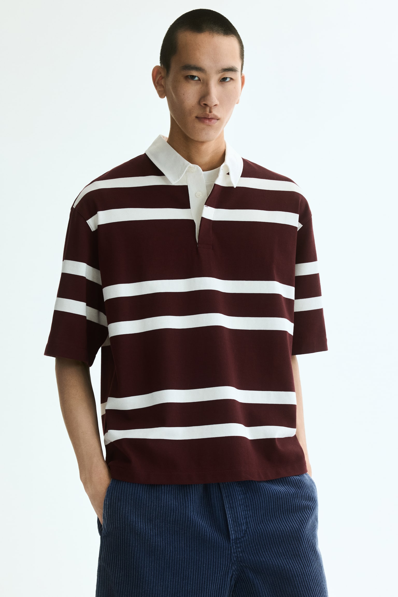 The polo shirt is displayed front-facing, showcasing broad horizontal stripes in maroon and off-white. It features an off-white turn-down collar, a two-button placket, dropped shoulders, and short sleeves, offering a relaxed fit. - 1