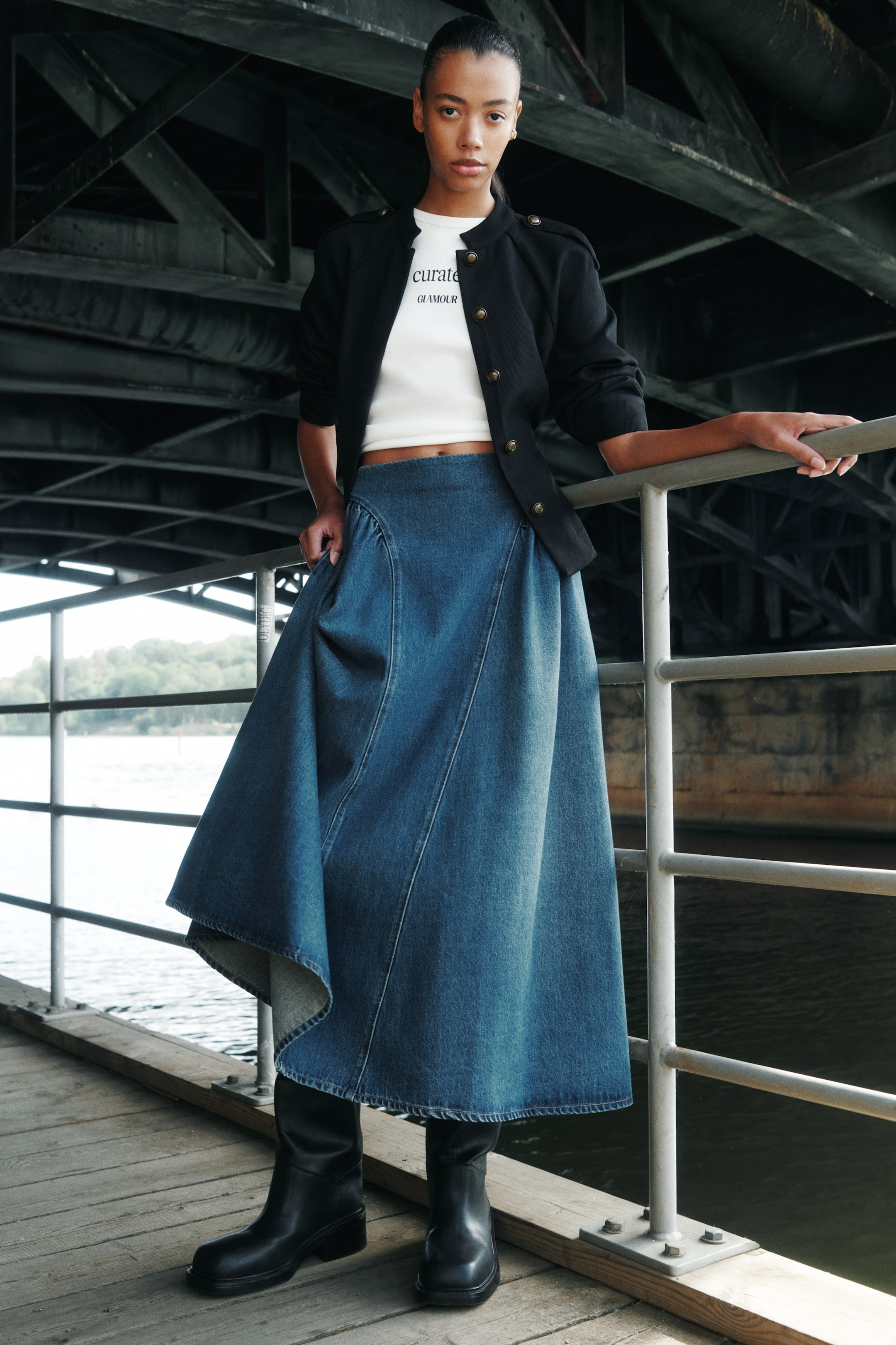 View larger image: A long A-line denim skirt in a blue wash is worn by the model, featuring a high waist and curved gathered seams at the hips that create volume. The skirt is styled with a cropped white T-shirt, a black buttoned jacket, and tall black boots.