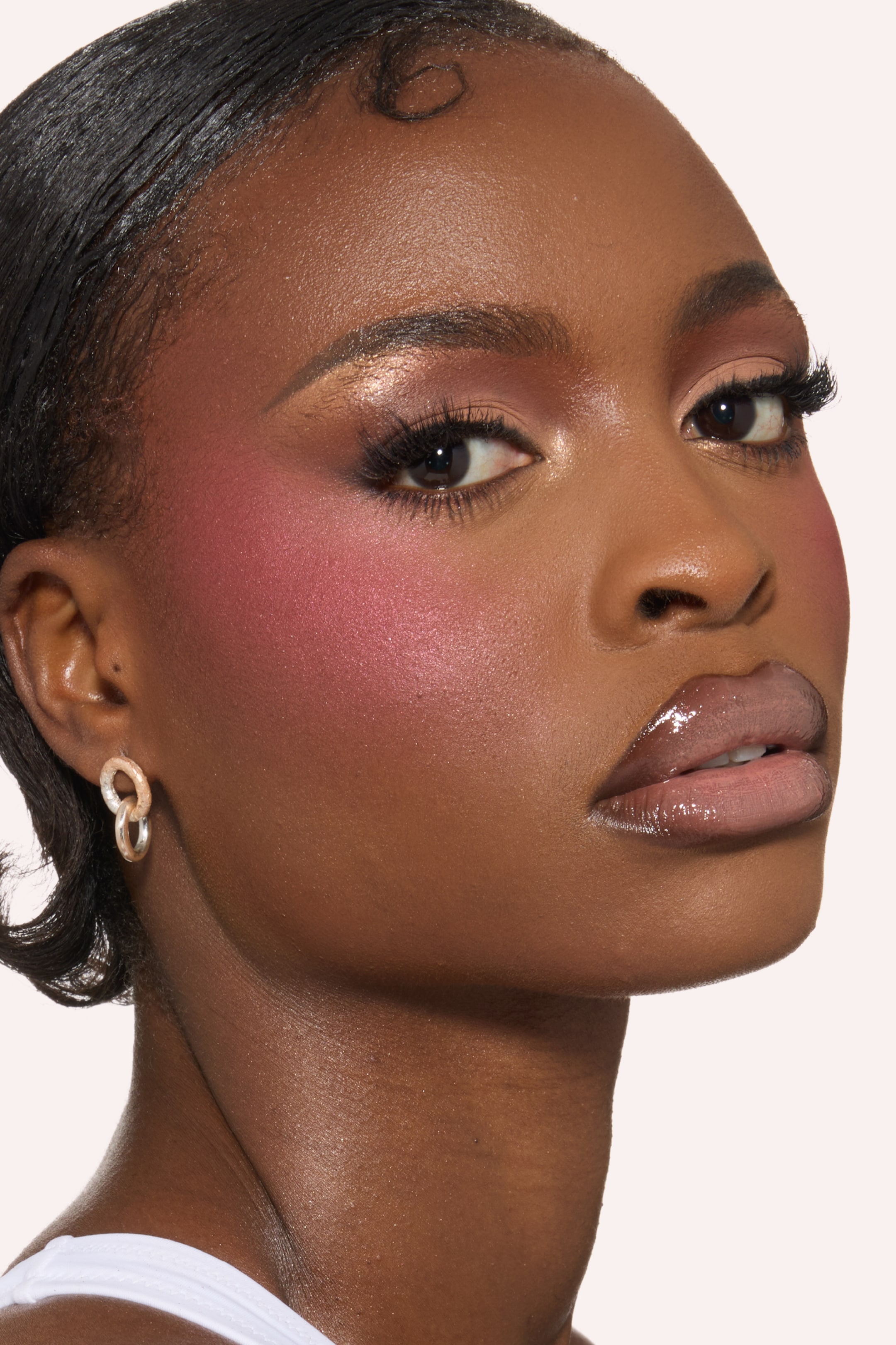 Vis større bilde: Rosa-rosa skinnende blush fremhever kinnbenet og tinningen, mens skinnende rosa og kobber øyenskygge pryder øyelokkene med definerte øyevipper. En glanset, nøytral rosa leppeglans dekker leppene, og huden viser en strålende finish.