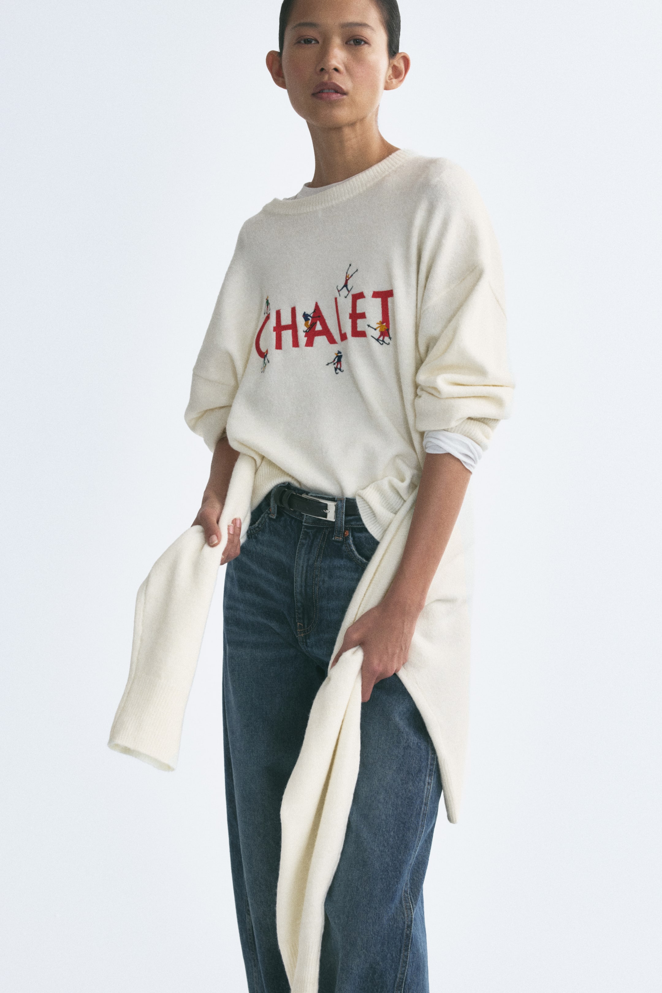 Ampliar la imagen: La modelo lleva un suéter tejido color crema de corte oversized, con cuello redondo acanalado, hombros caídos y un gráfico bordado en rojo en el pecho. La modelo sostiene los extremos drapeados de una prenda tejida a juego, anudada a la cintura, combinada con jeans azules y un cinturón negro.
