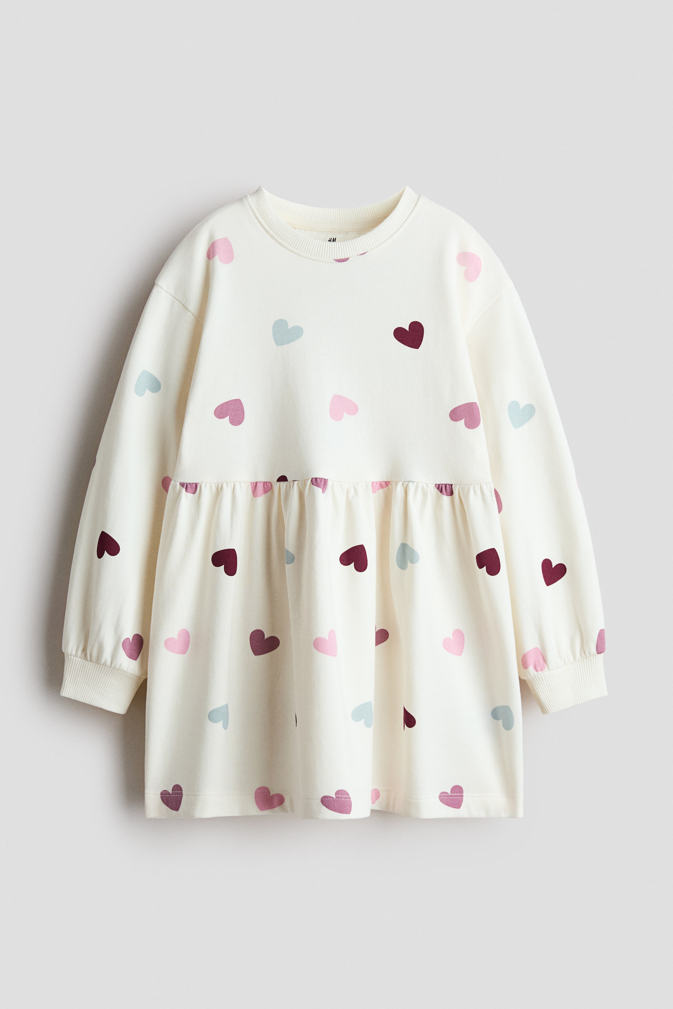 View larger image: An oversized, off-white fleece dress is shown from the front, featuring an allover pattern of small hearts in shades of pink, burgundy, and light blue. The dress is designed with a round rib-trim neckline, dropped shoulders, long sleeves with ribbed cuffs, and a gathered seam at the waist that creates a flared skirt.
