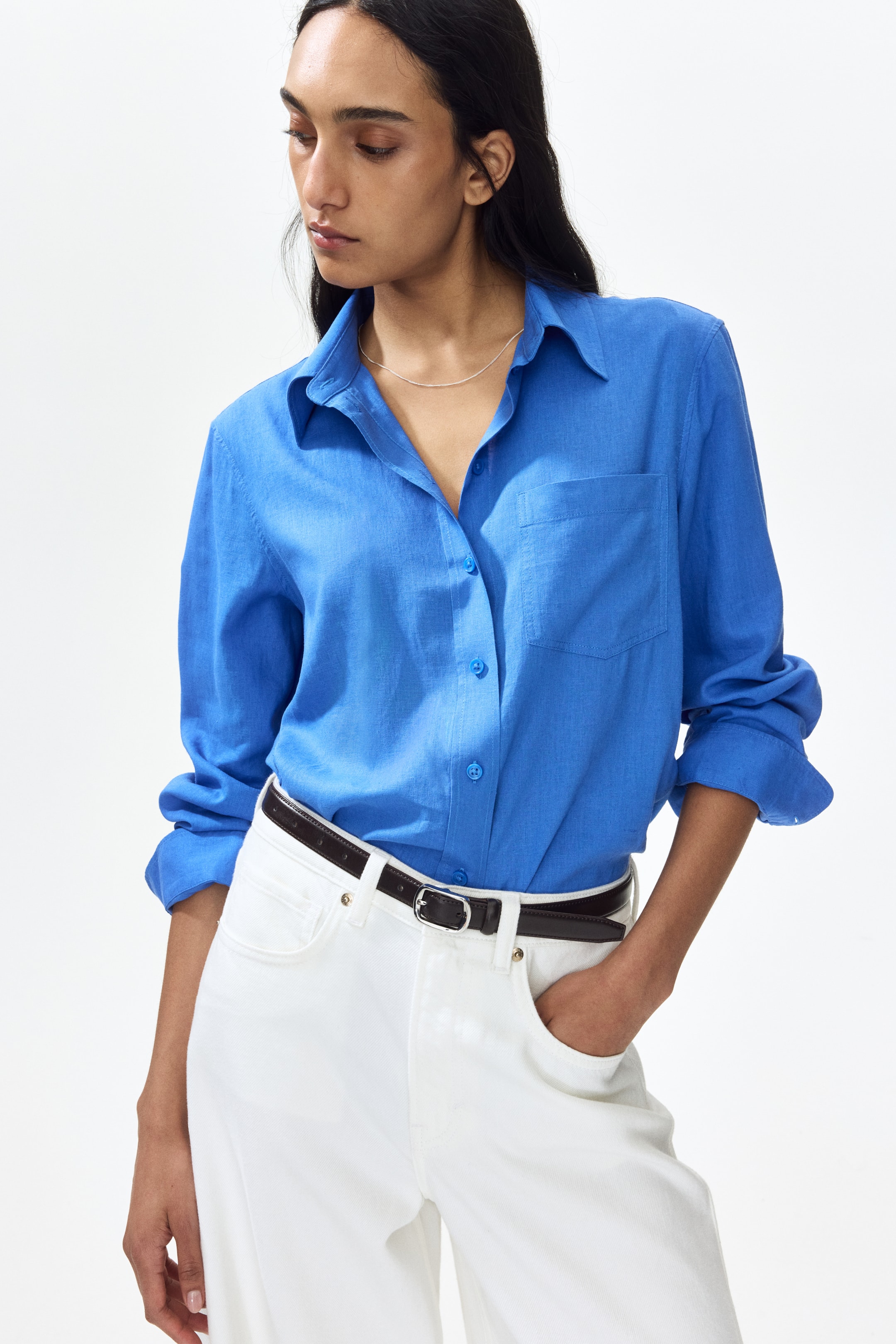 View larger image: A blue long-sleeved shirt is showcased from the front, slightly angled to the left, featuring a textured fabric, a pointed collar, and a patch pocket on the left chest. It is buttoned down the front, with its sleeves rolled up at the cuffs, and a silver necklace is visible in the open collar.