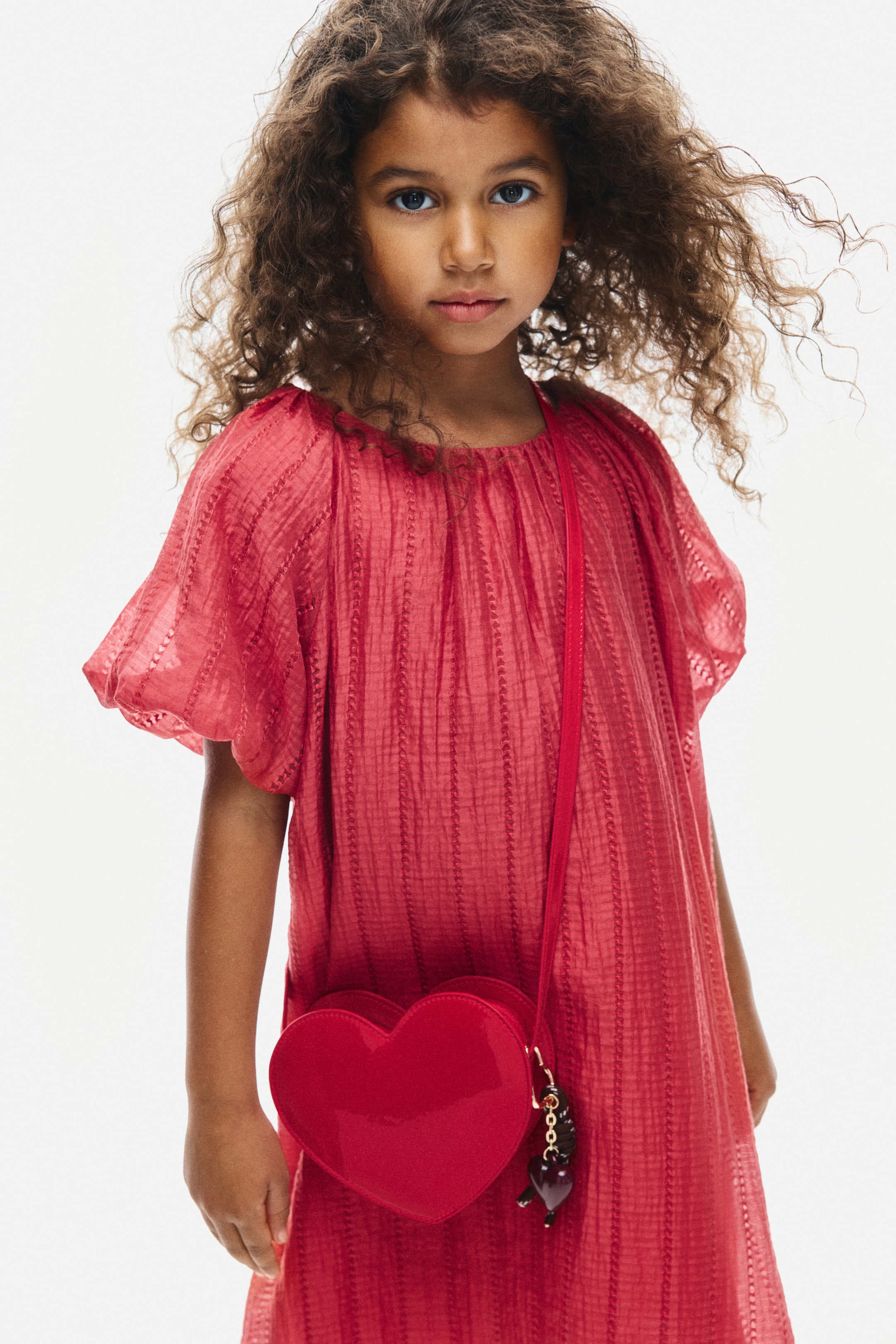 View larger image: A red A-line dress in a soft, textured weave is shown from the front, featuring a gathered round neckline and short, voluminous raglan sleeves with elasticated cuffs. A red, heart-shaped crossbody bag is draped over the front of the dress’s loose silhouette.