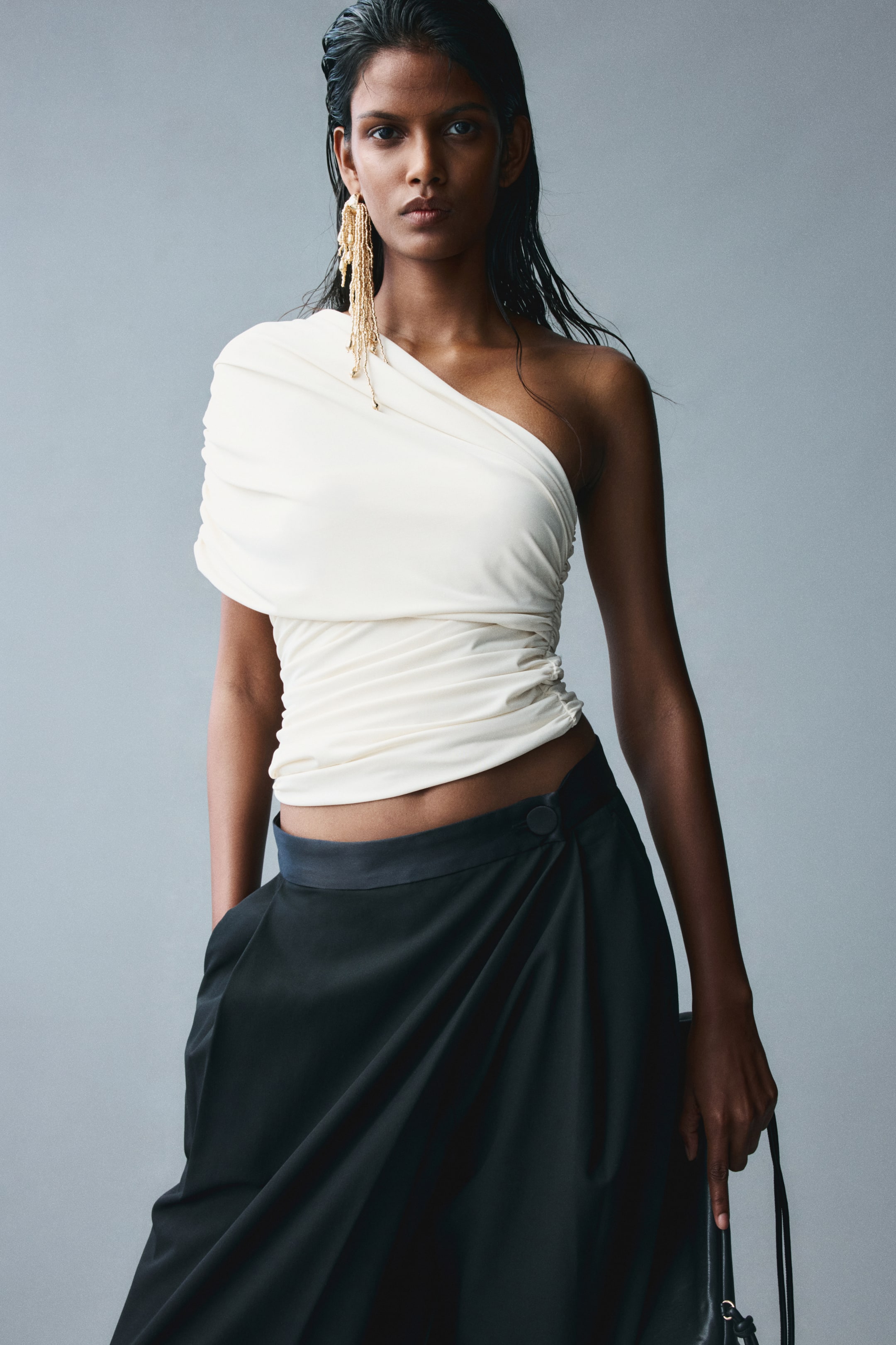 View larger image: A cream-coloured, one-shoulder cropped top featuring a diagonal neckline and a draped sleeve panel on one side is being worn by the model. The fitted top has gathered seams creating a ruched effect and is paired with low-rise black trousers.