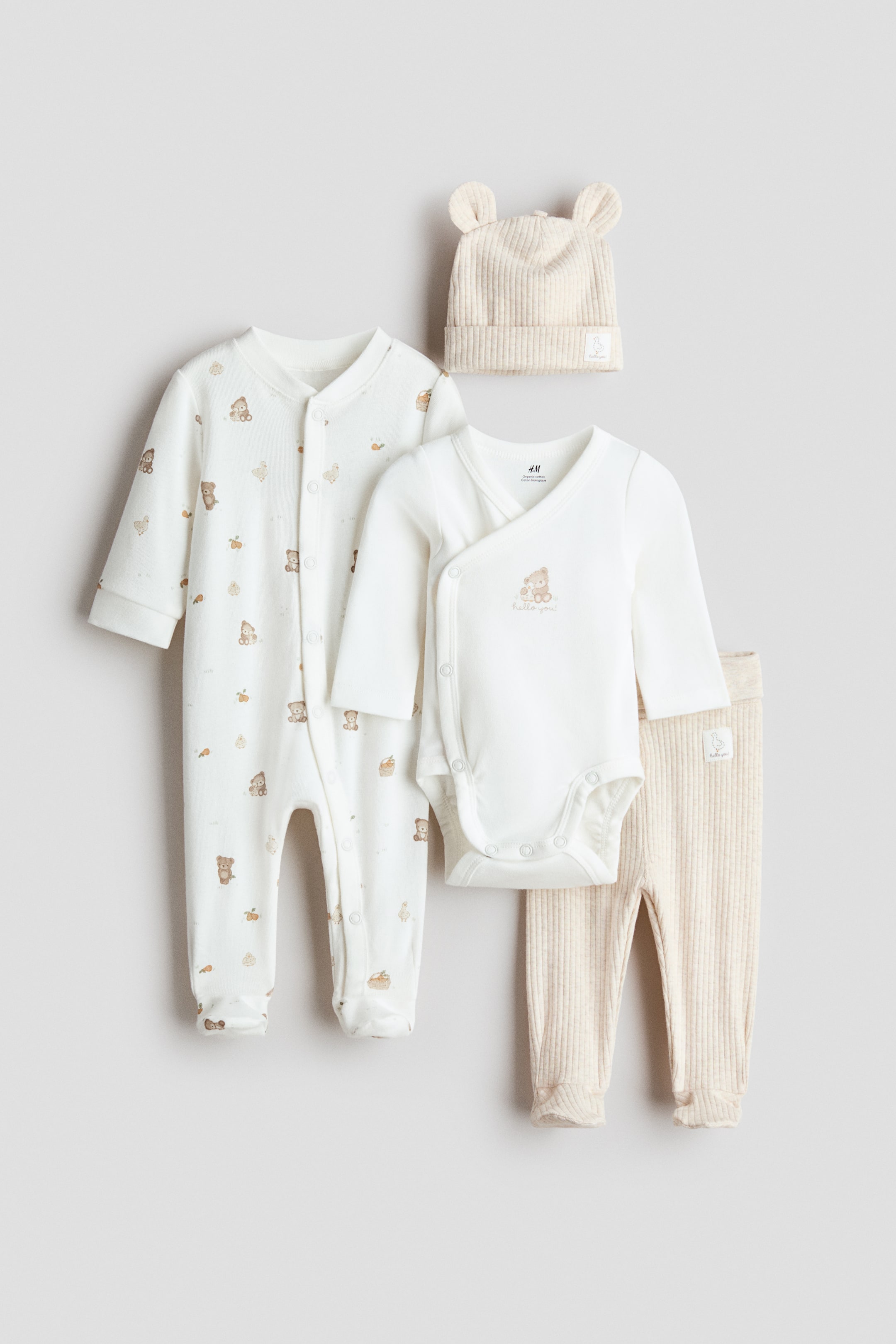 View larger image: A four-piece baby clothing set is being presented laid flat, comprising a white footed sleepsuit featuring an all-over brown bear and motif pattern with a full-length snap closure. A white long-sleeve wrap-style bodysuit is also showing an embroidered bear and "hello you" text, along with light beige ribbed trousers with ribbed cuffs, and a light beige ribbed hat with ear-like protrusions.