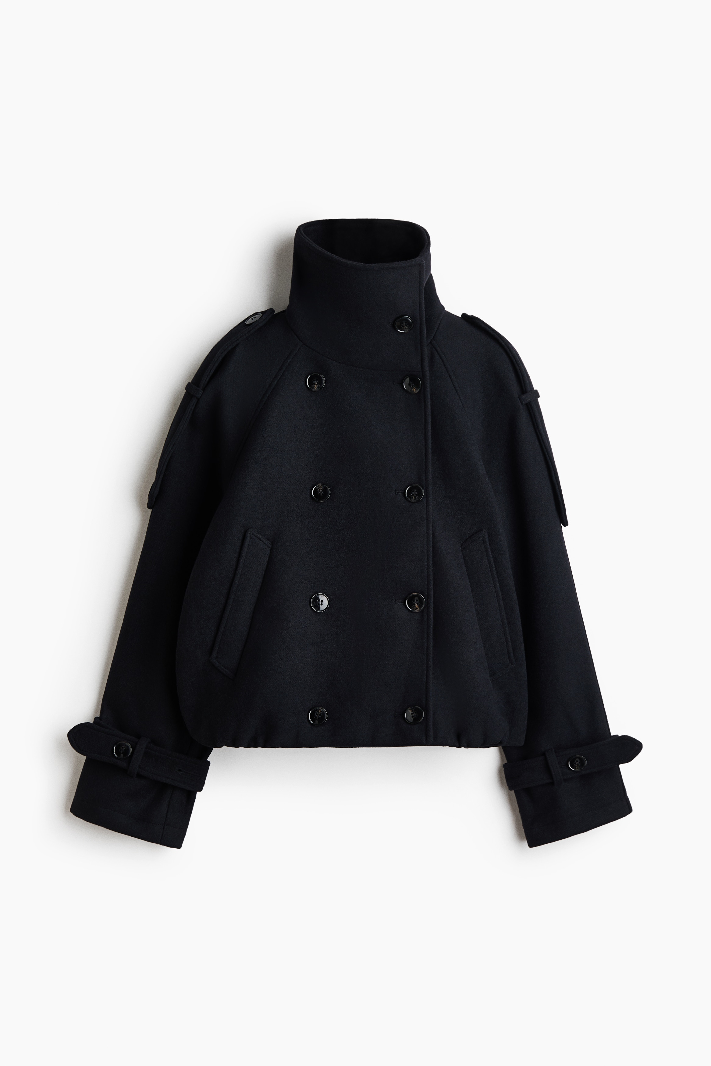 A modelo veste um casaco curto, de abotoamento duplo, em tecido feltro escuro. O casaco oversized apresenta gola alta tipo funil com botões, fecho frontal cruzado de abotoamento duplo e punhos com patilhas abotoadas, sendo combinado com calças em pele escura e uma alça fina ao ombro que cruza o peito.