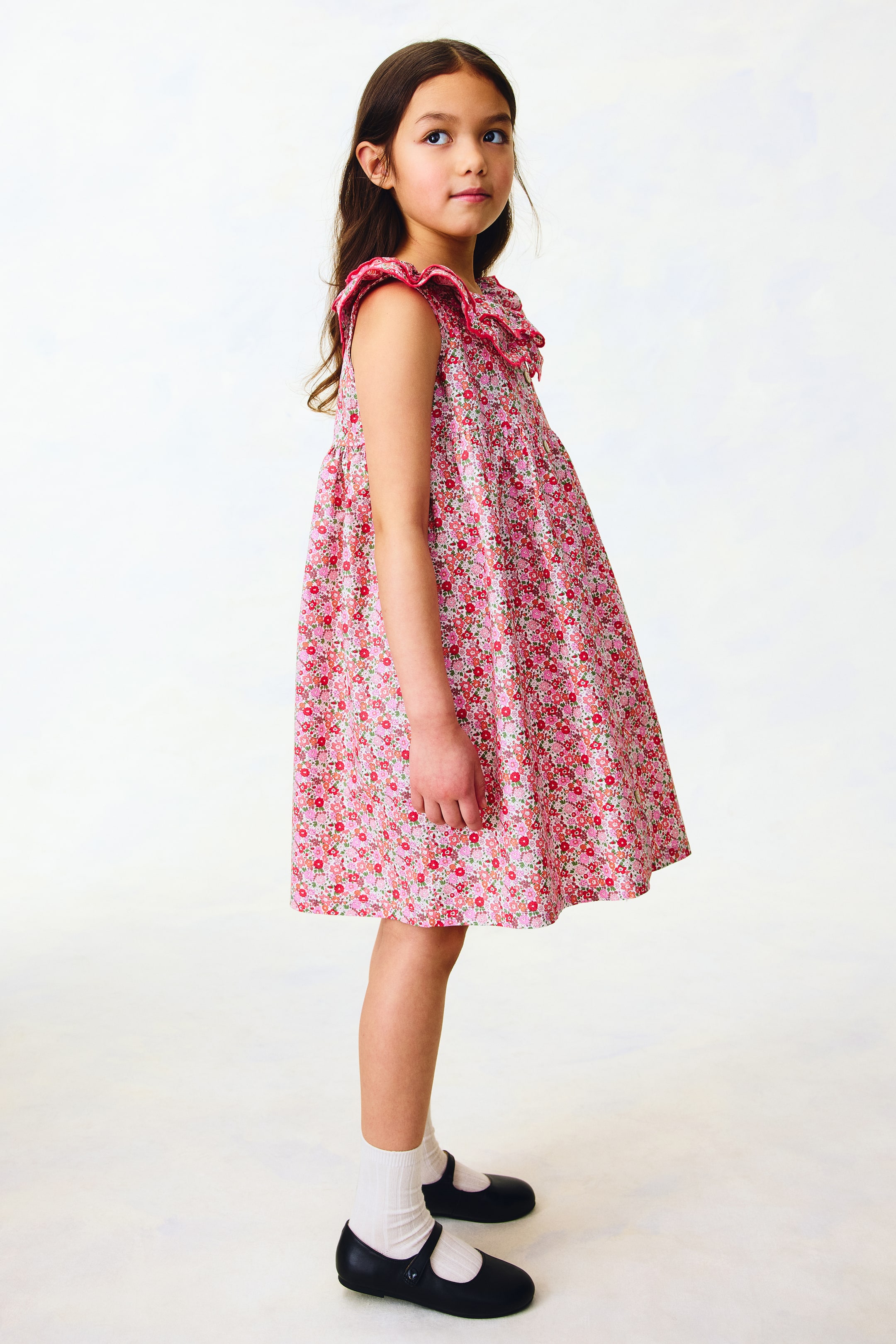 Agrandir l'image: Une robe florale rose pour enfant présente un col froncé avec une garniture à volants dans une teinte de rose plus foncée, et elle arbore une silhouette évasée sans manches en ligne A, tournée vers la droite.