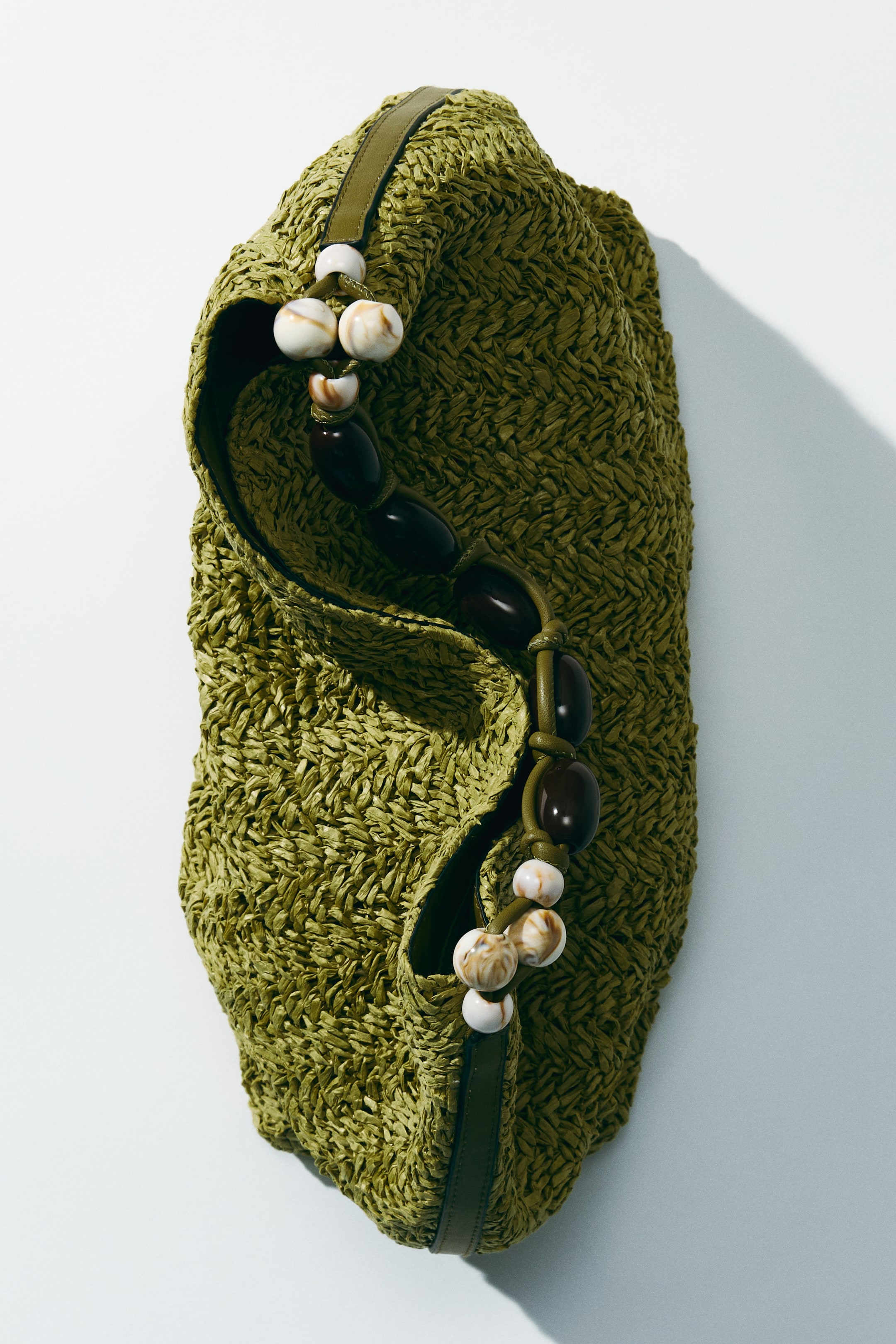 View larger image: A shoulder bag in olive green is displayed from a slightly raised angle, showcasing its braided paper straw design. The handle, adorned with dark brown oval beads and smaller cream marbled beads, elegantly arches over the top opening.