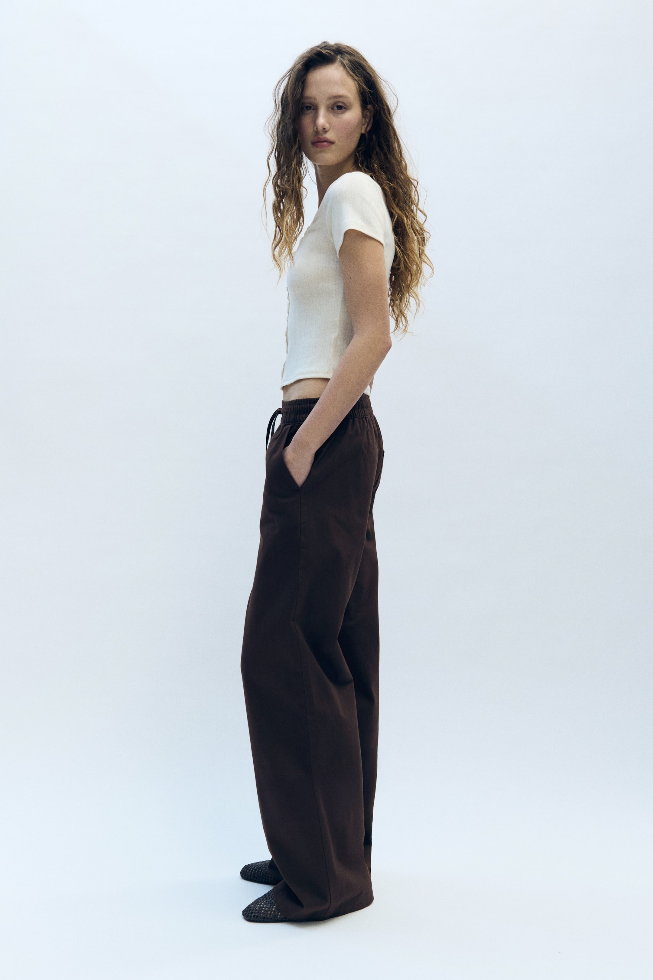 View larger image: An off-white ribbed knit short-sleeve top with a button-front placket is showcasing a slim, cropped fit from the side. It is paired with dark brown wide-leg pants, featuring an elasticated drawstring waist and presenting a relaxed, full-length silhouette, elegantly pooling over dark woven flat shoes.