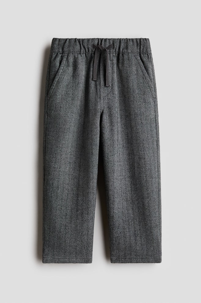 These dark gray herringbone trousers are facing front, featuring an elasticated waistband with an adjustable drawstring and side slant pockets. The trousers are falling into a relaxed, straight-leg fit in a textured woven fabric. - 1