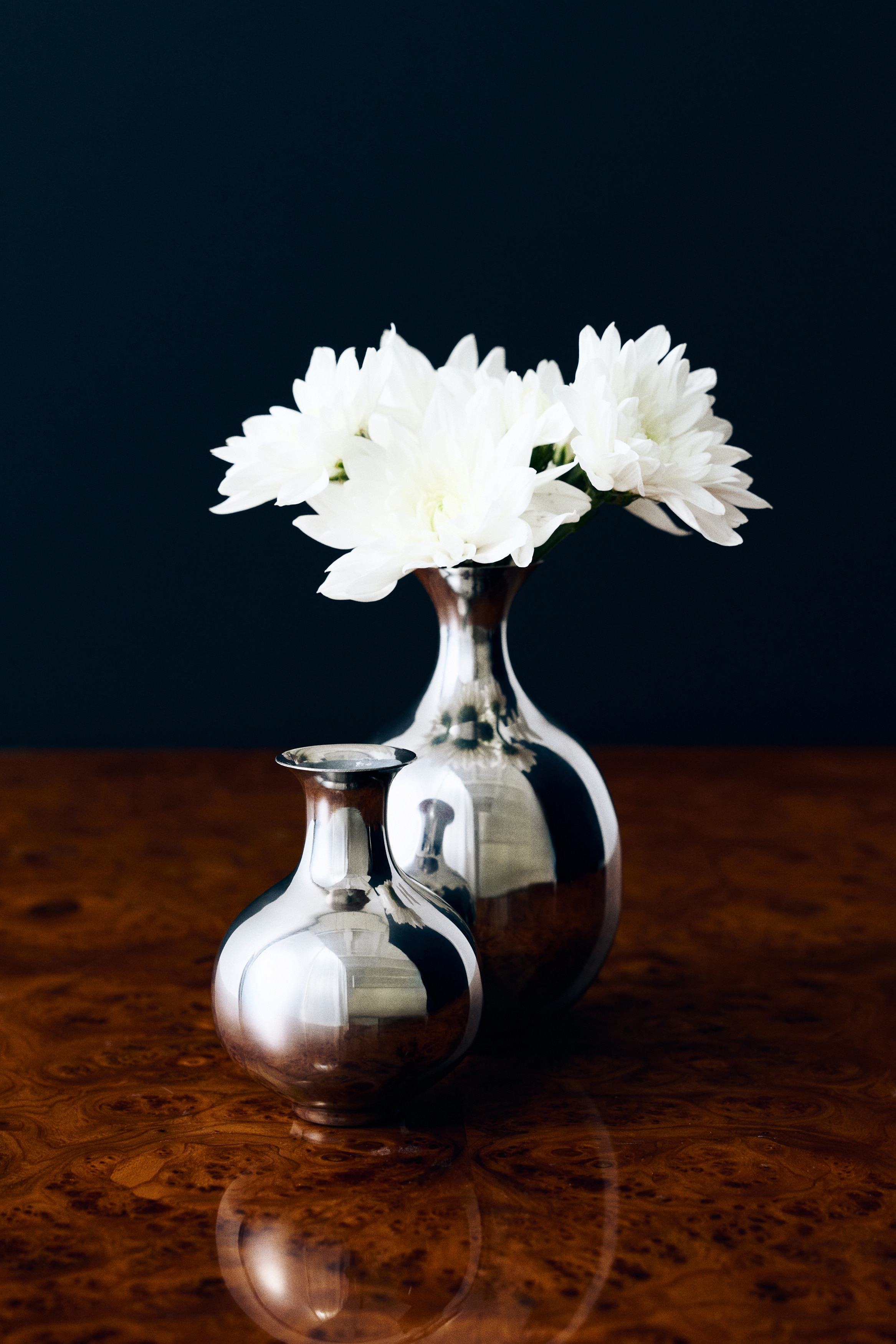 A polished silver-toned metal vase is standing facing front, showcasing a bulbous body, a narrow neck, and a flared rim. The highly reflective, metallic surface is displaying dark, mirrored reflections across its curved shape.