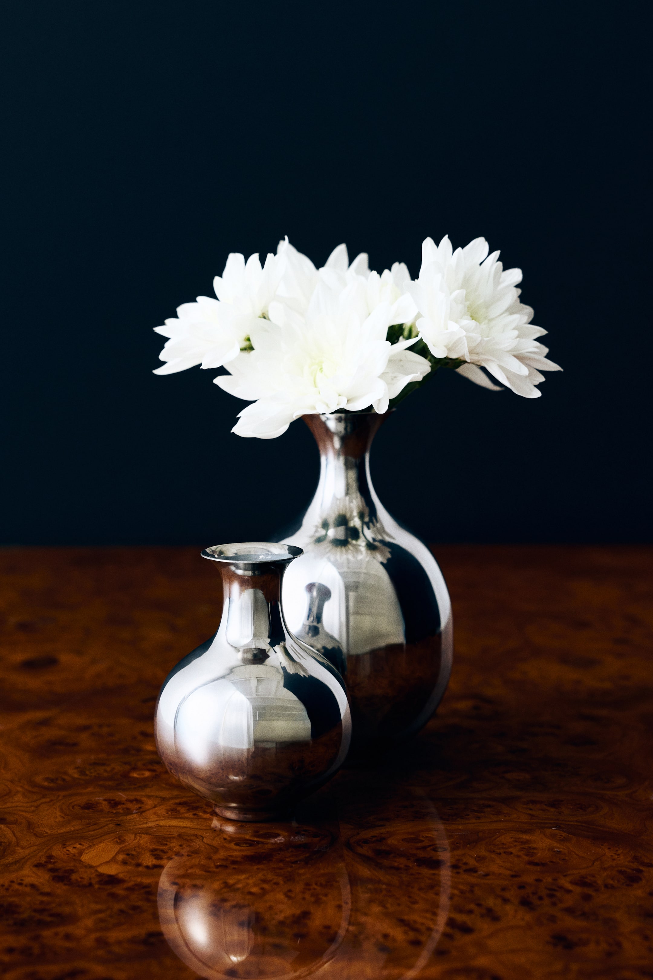 View larger image: A small stainless steel vase with a rounded, bulbous body, a narrow neck, and a flared rim is placed facing the front, with its highly polished surface reflecting the surroundings. A larger matching stainless steel vase holding white flowers stands directly behind it, highlighting the product's scale and reflective design.