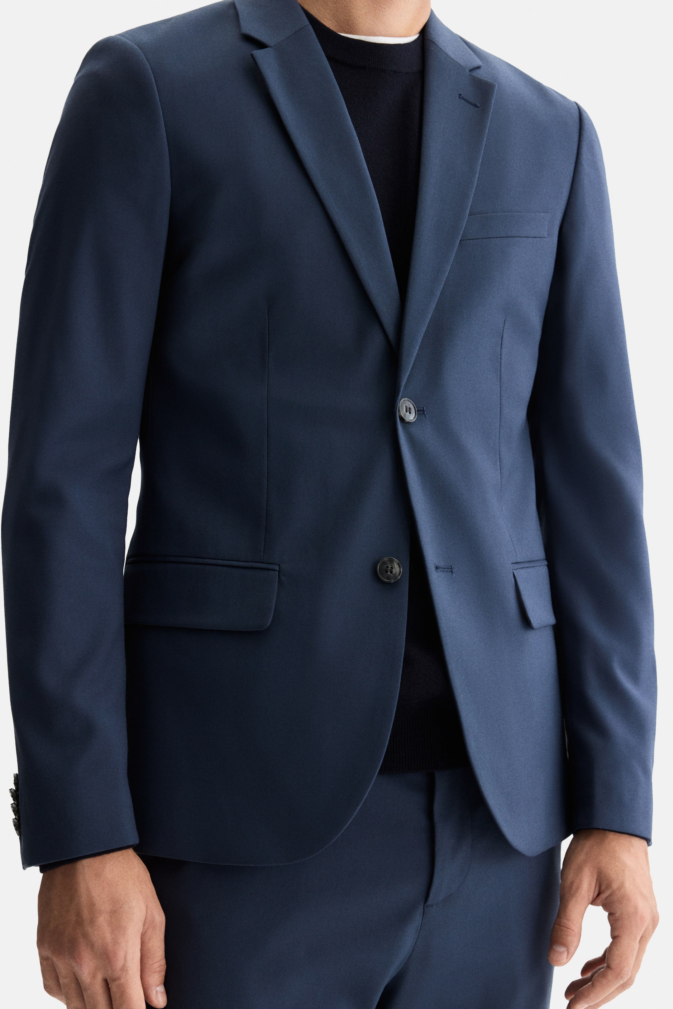 Ampliar la imagen: Una chaqueta de traje azul marino y pantalones a juego están siendo usados por el modelo. La chaqueta presenta una solapa con muesca, un frente de dos botones con el botón superior abrochado y bolsillos con solapa. Debajo se lleva un suéter oscuro de cuello redondo, con una prenda blanca visible en la línea del cuello.