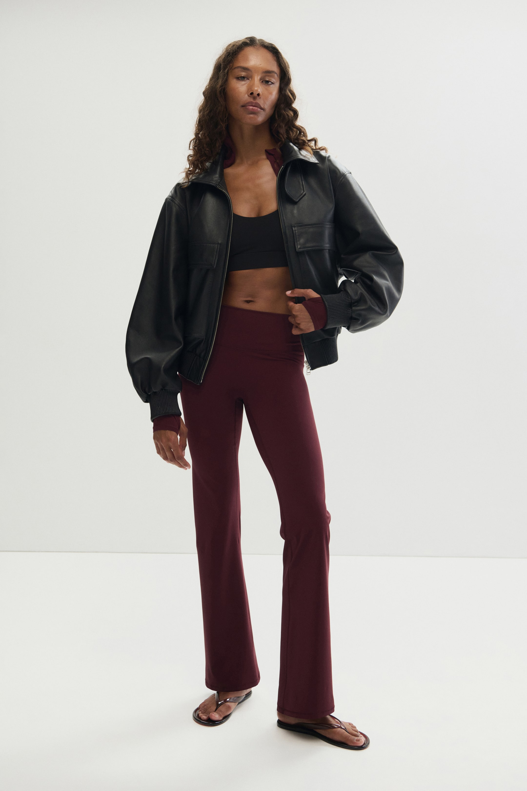 View larger image: SculptMove? The model is wearing burgundy flared sports leggings featuring a high waist and a flared silhouette. She is holding the waistband, while a matching burgundy long-sleeved top is layered under a black jacket.