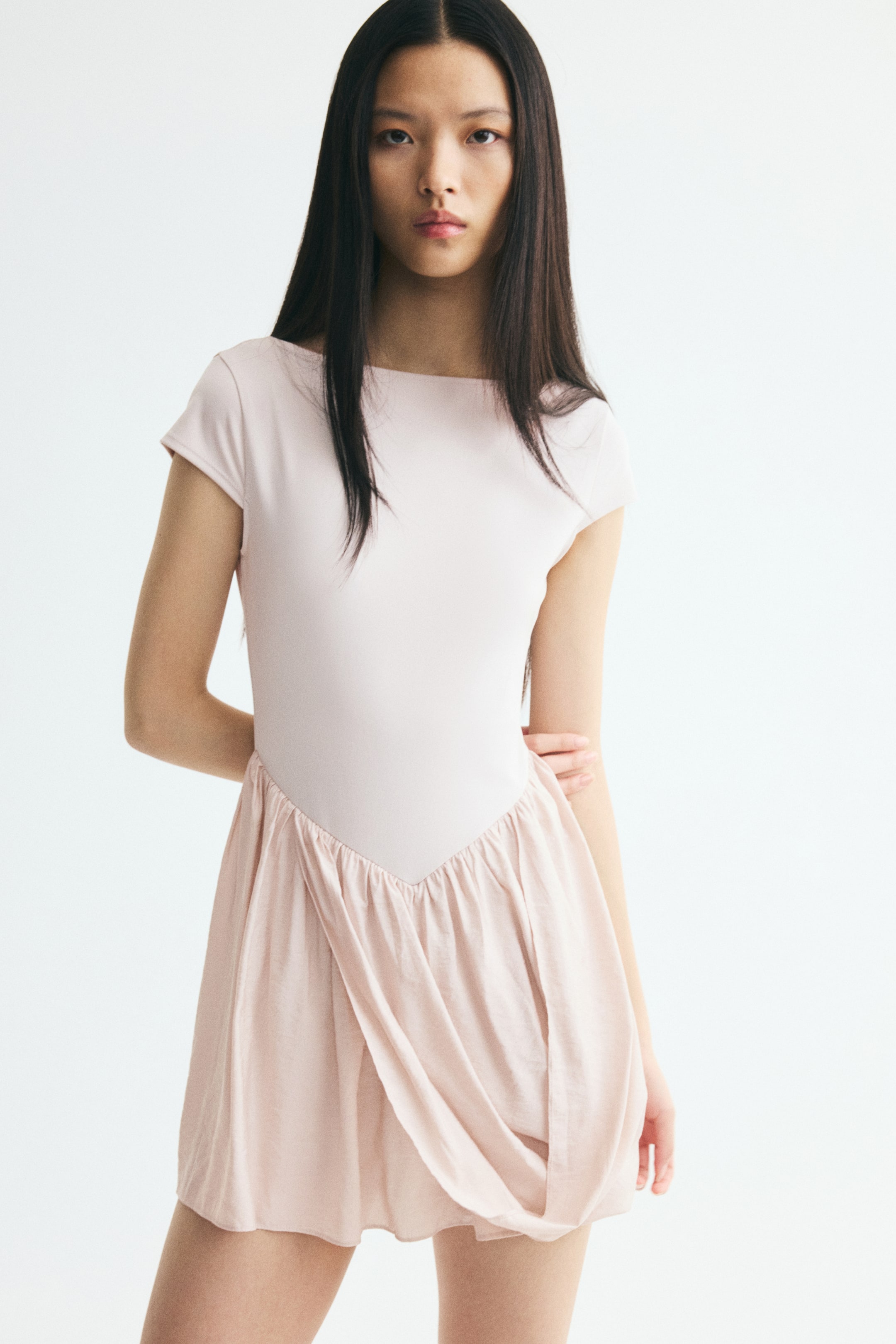 View larger image: The model is wearing a light pink mini dress, showcasing a fitted bodice with a boat neckline and cap sleeves. The dress features a gathered V-shaped waistline and a draped front panel on the flared skirt.