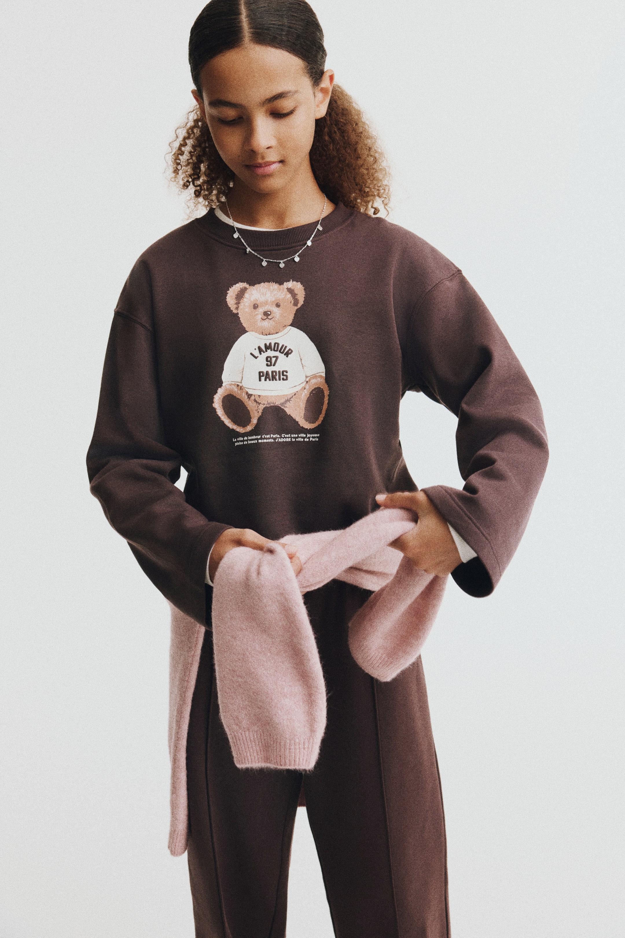 View larger image: This dark brown fleece sweater and matching track pants set is shown from the front. The sweater features a ribbed crew neck, dropped shoulders, and a printed teddy bear graphic on the front, while the matching straight-leg track pants have sewn-in creases down the centre of each leg as a light pink knit sweater is tied around the waist.
